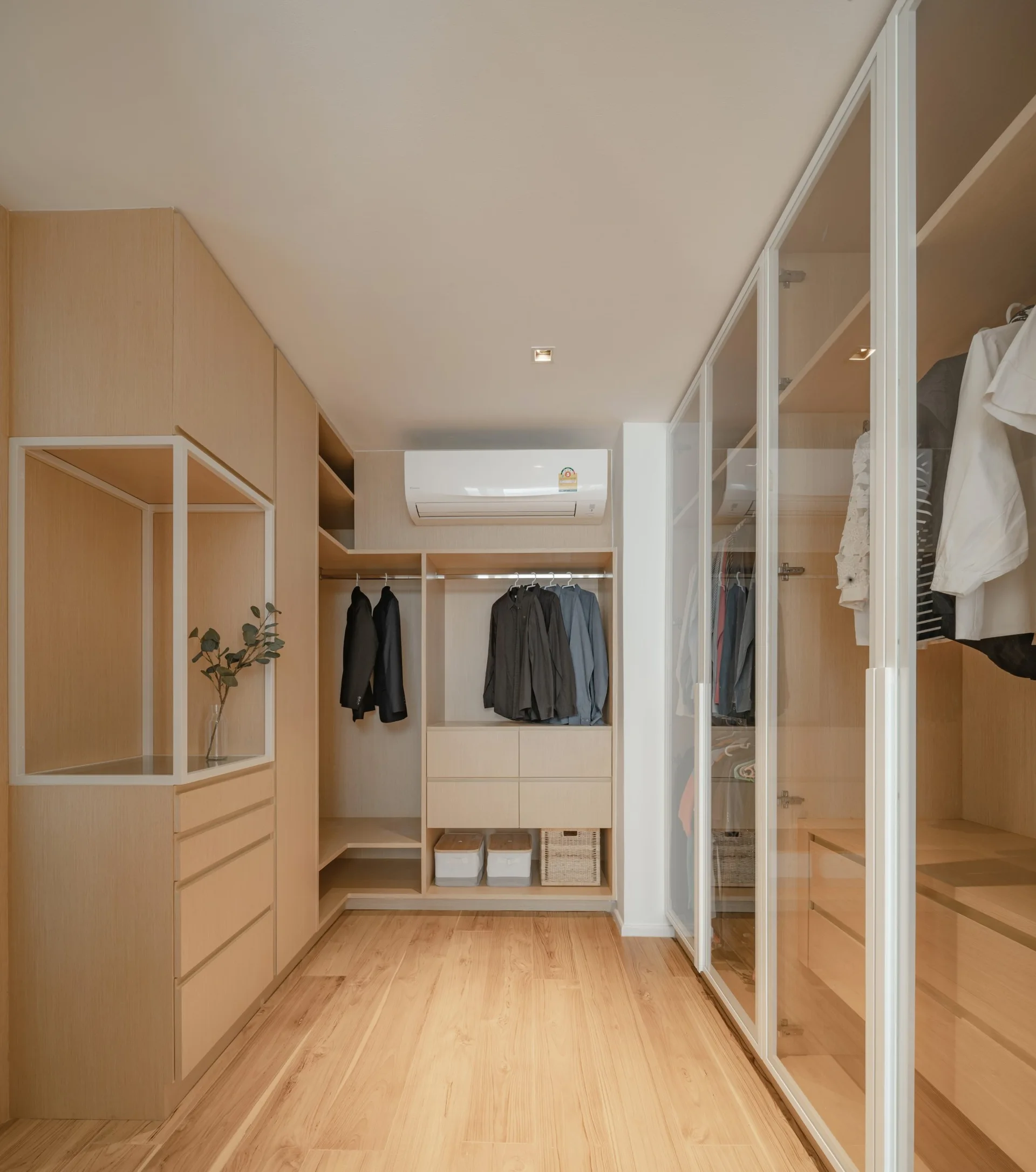 A walk-in closet features custom light wood cabinetry, integrated drawers, hanging clothes, and glass-front wardrobes over wood floors.
