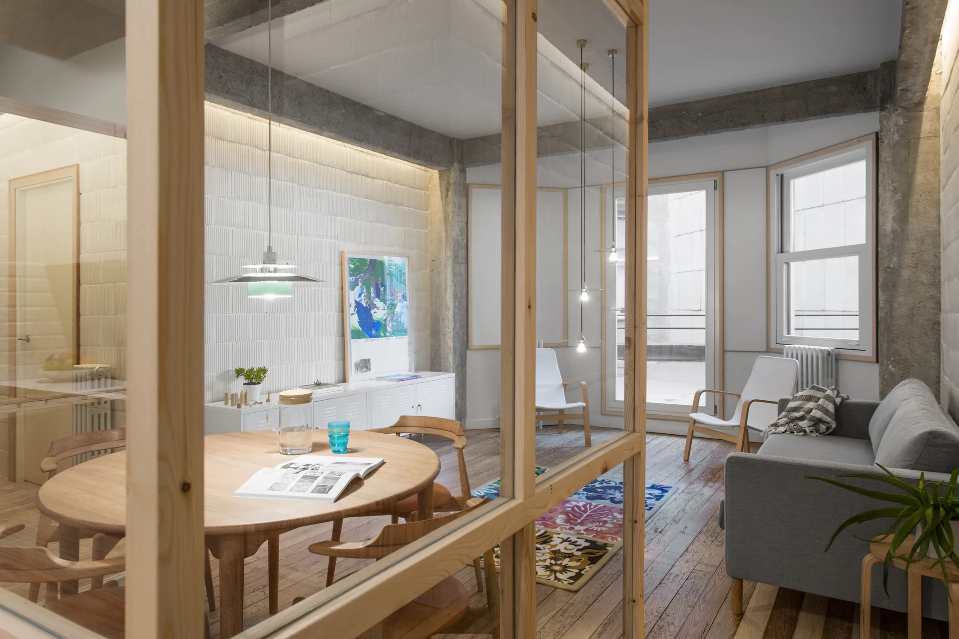 A modern living and dining space featuring a round wood dining table, a grey sofa, and a wood-framed glass partition in the foreground.