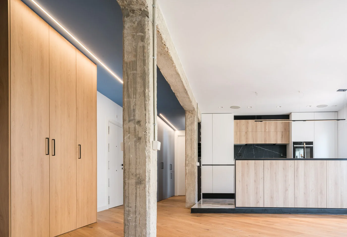 A modern open-plan kitchen featuring light wood cabinets, an exposed concrete column, and a dark blue ceiling section with LED lighting.