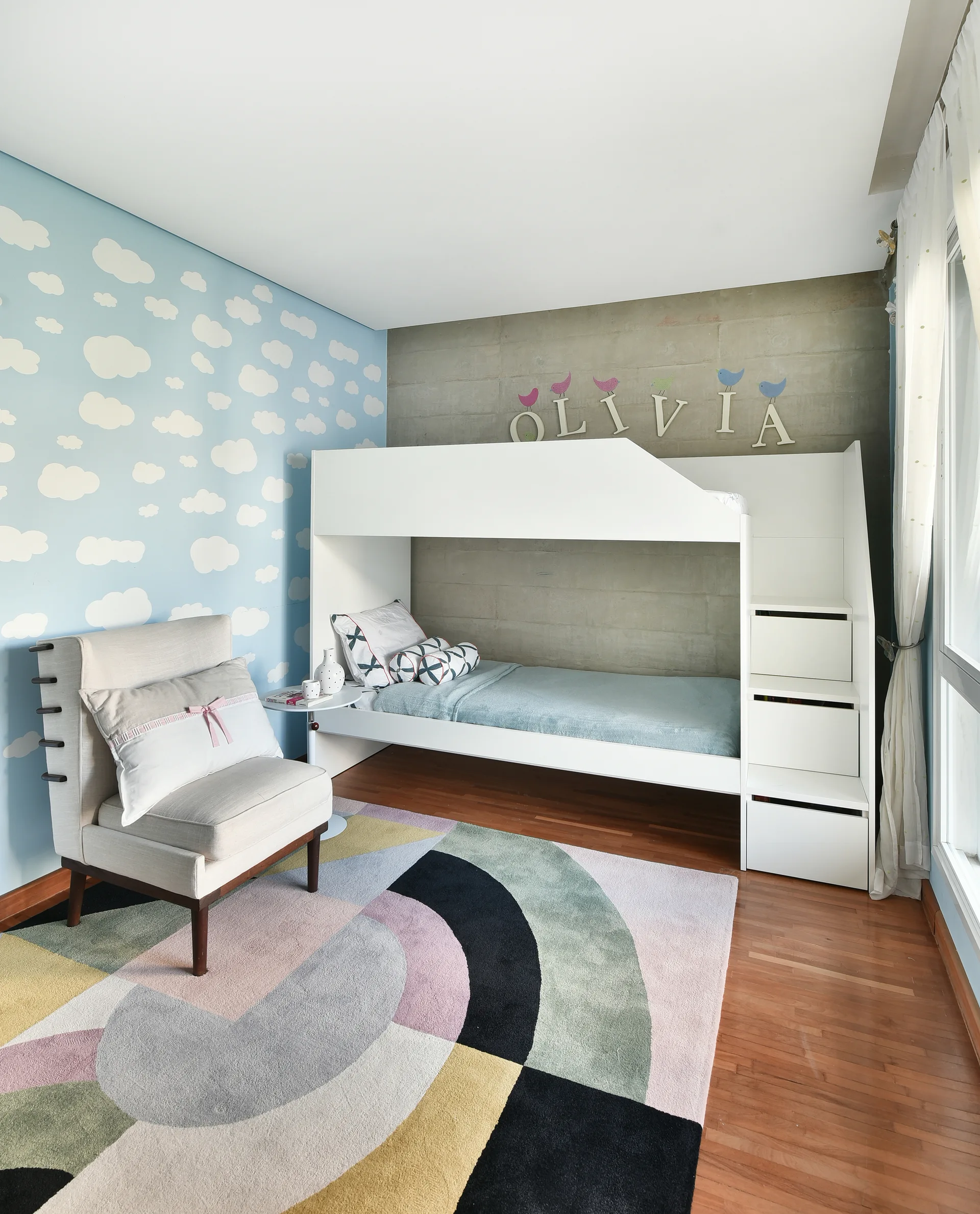 A child's bedroom features a white wooden bunk bed against a concrete wall, blue cloud-patterned wallpaper, and a colorful geometric area rug.