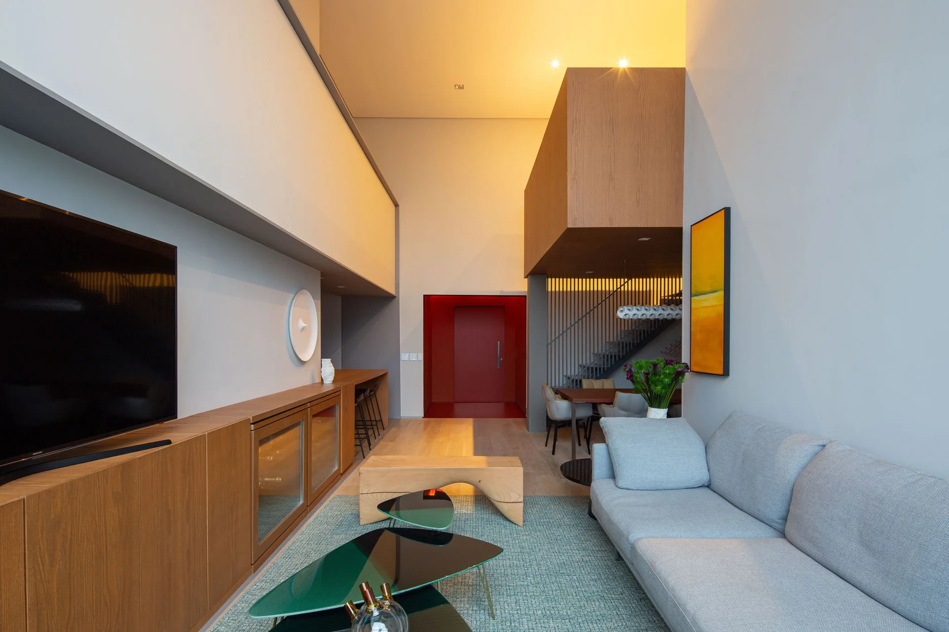 A contemporary living room features a grey sofa, a long wooden media console, a double-height ceiling, and a bright red doorway in the background.