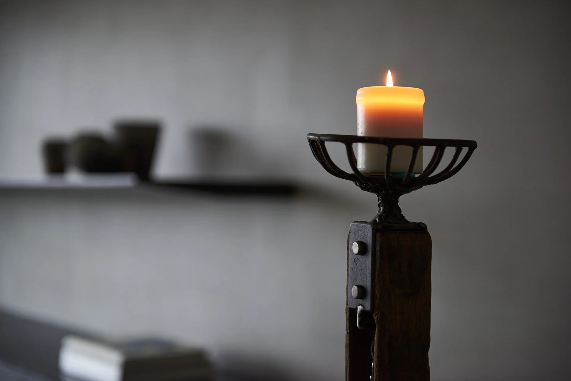 A close-up of a lit amber pillar candle sitting in an industrial metal and wood candlestick with a blurred grey wall in the background.