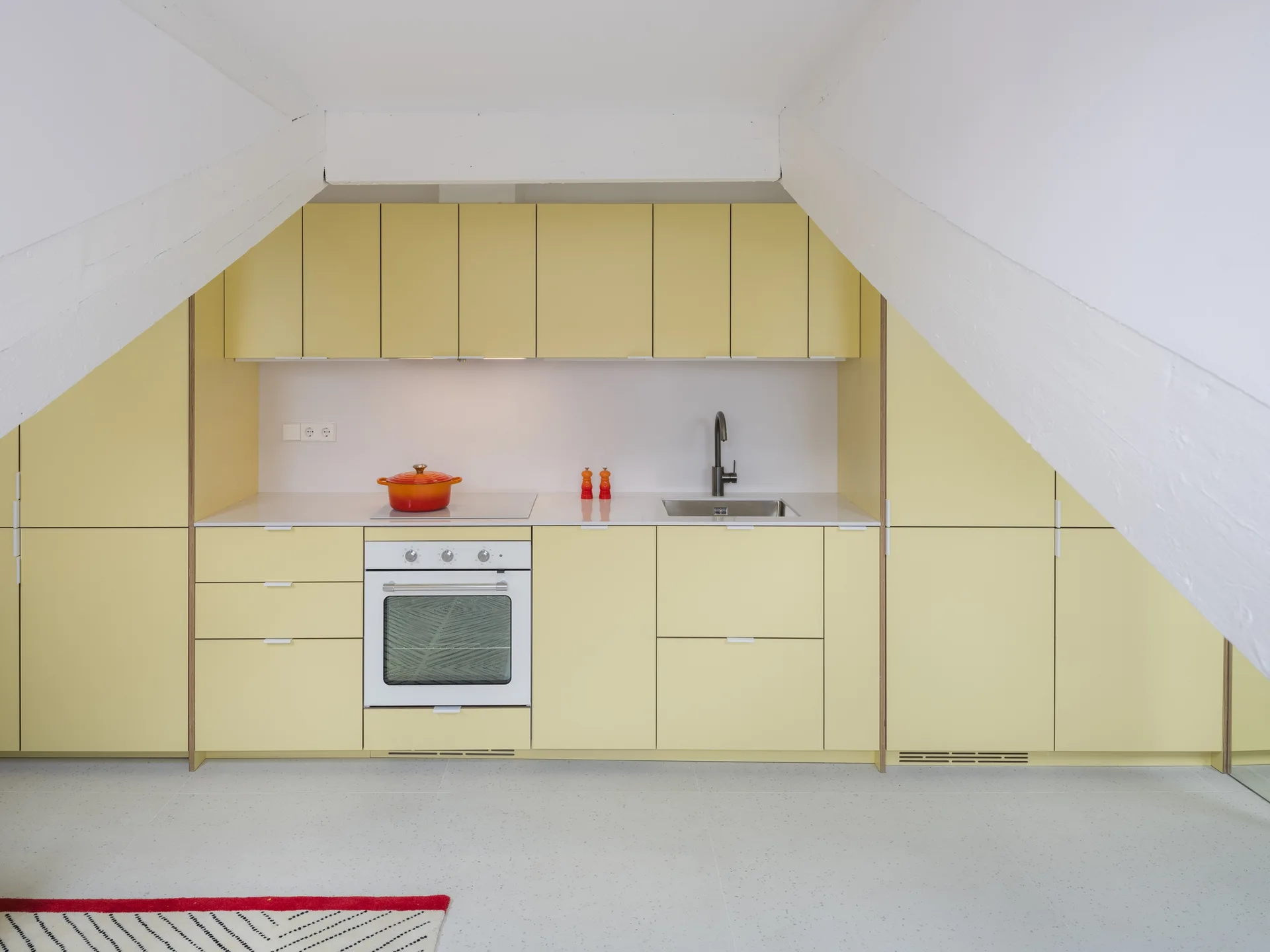 A minimalist kitchen set under slanted white attic beams featuring pale yellow cabinetry, a white built-in oven, and a white countertop.