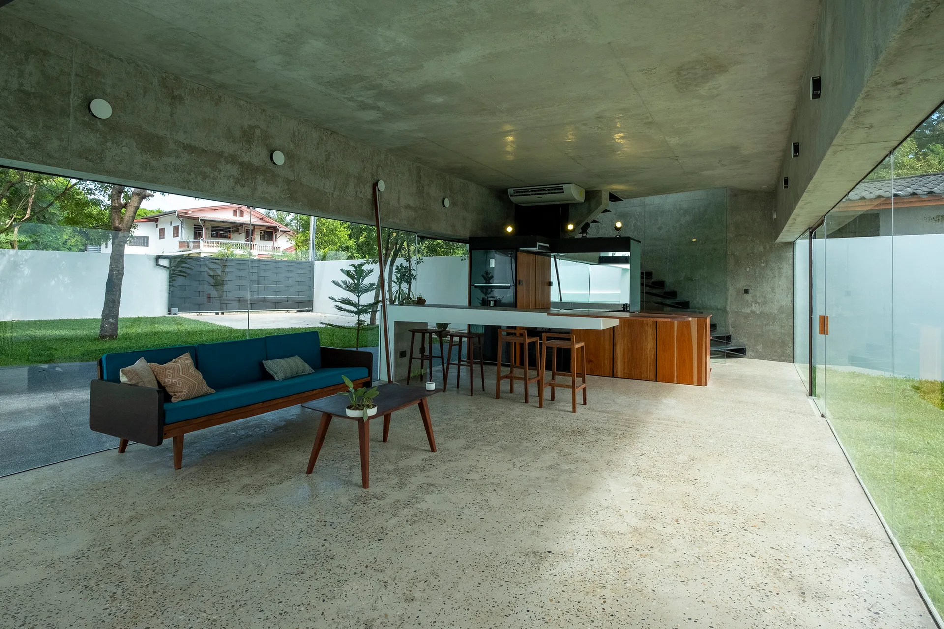 A modern open-plan room features exposed concrete walls and ceiling, a blue sofa, and glass walls looking out to a green lawn.