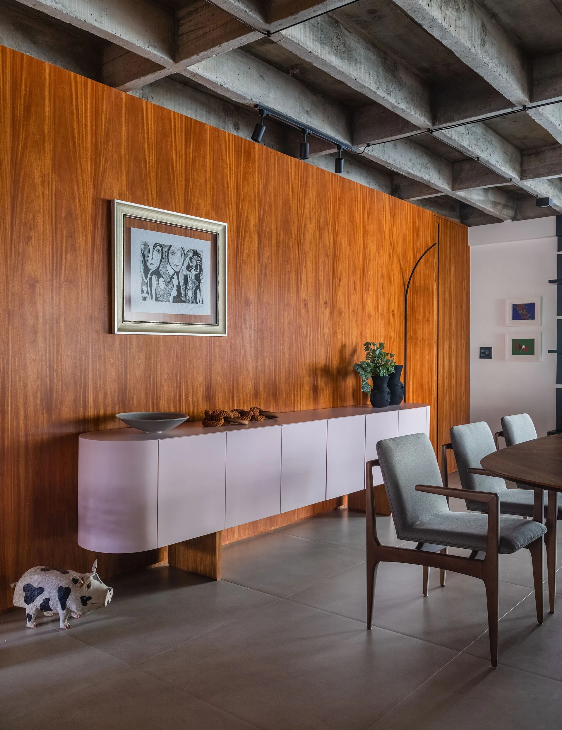 A dining room features a curved pink sideboard against a wood-paneled wall beneath an exposed concrete waffle ceiling.