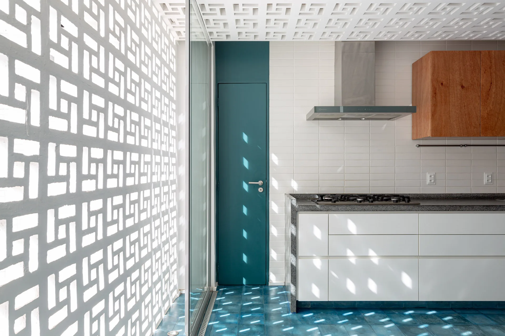 A modern kitchen featuring white cabinets, a wooden upper cupboard, blue floor tiles, and a white geometric breeze block wall filtering natural light.
