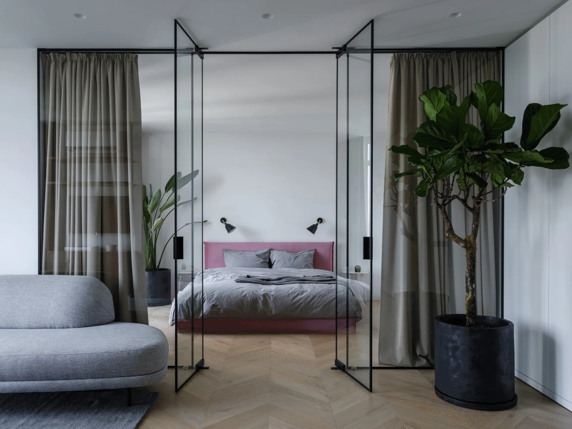 A modern bedroom with a pink bed frame is visible through a black-framed glass partition wall, featuring chevron wood floors and a grey sofa in front.