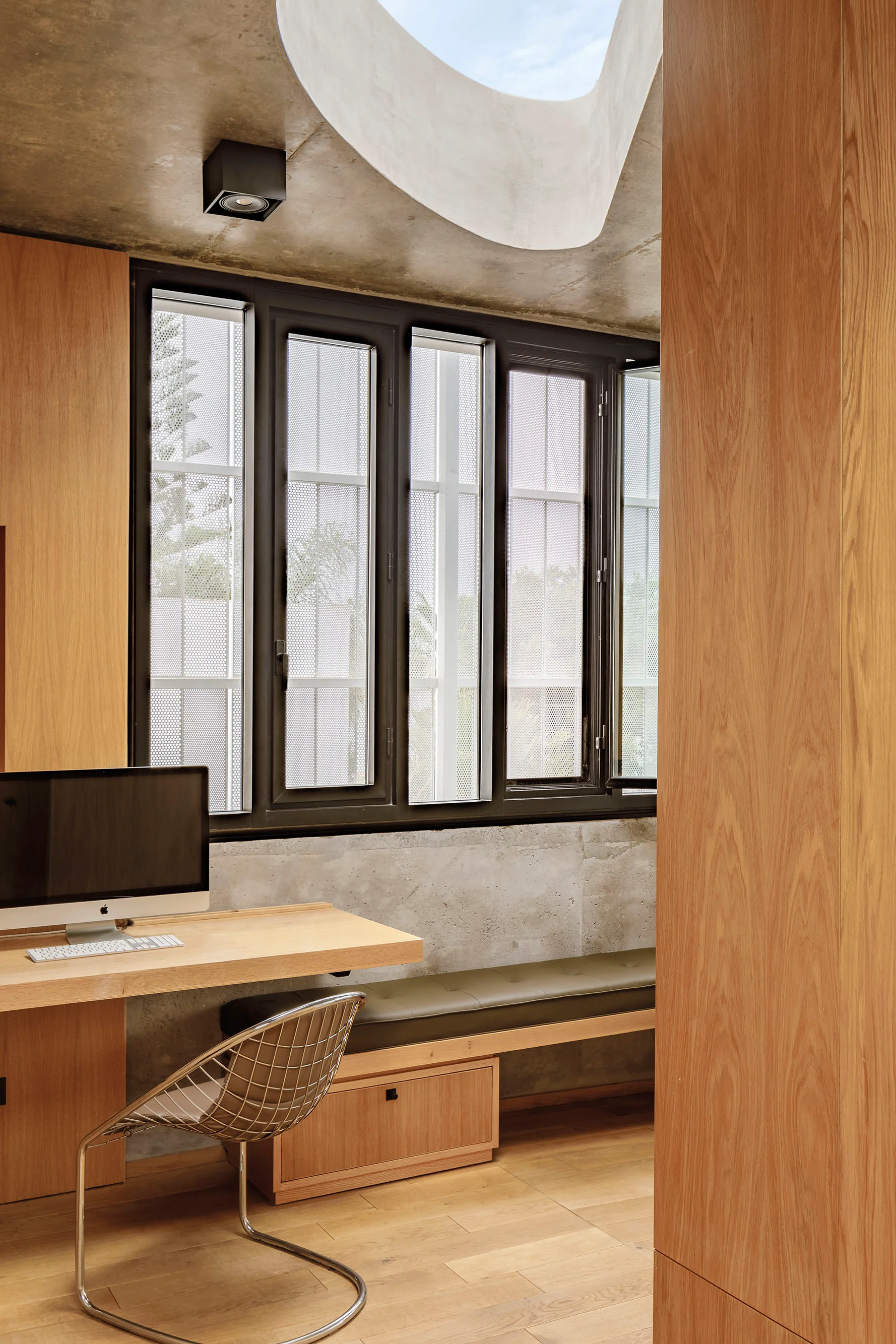 A modern home office with a wooden built-in desk, a metal grid chair, an olive bench, and a circular skylight in the concrete ceiling.