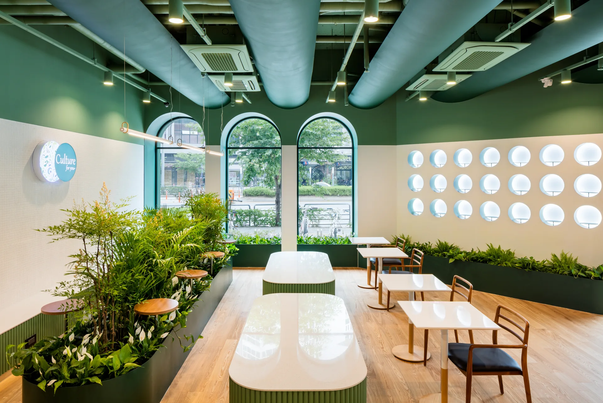 A modern cafe interior features white tables, wood chairs, built-in floor planters with leafy greenery, and exposed green ceiling ducts.