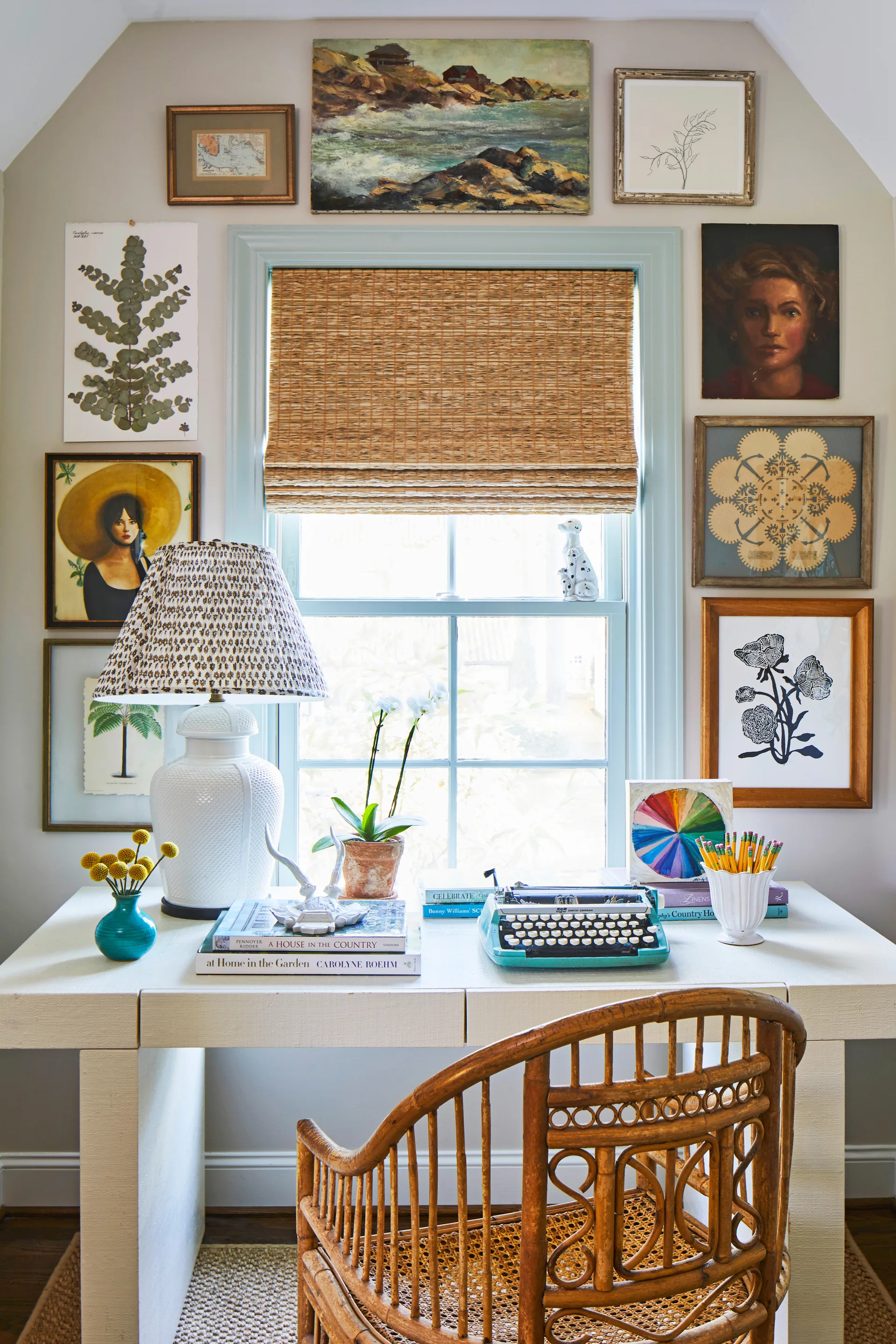 An off-white writing desk with a turquoise typewriter, a white ceramic lamp, and a rattan chair in front of a window wrapped in a gallery wall.