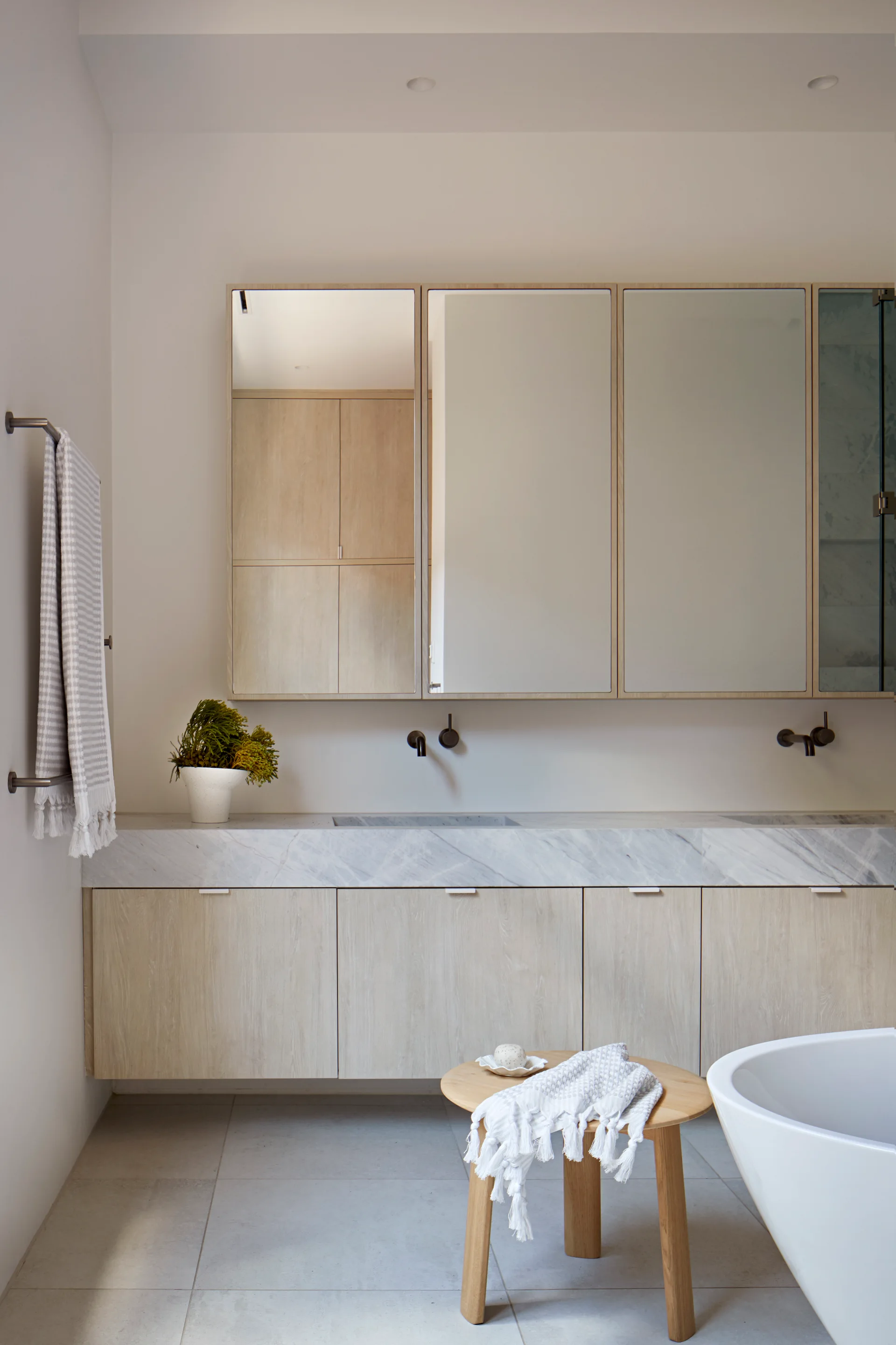A minimalist bathroom featuring a floating light wood vanity, marble countertop, wall-mounted dark faucets, and a round wooden stool.