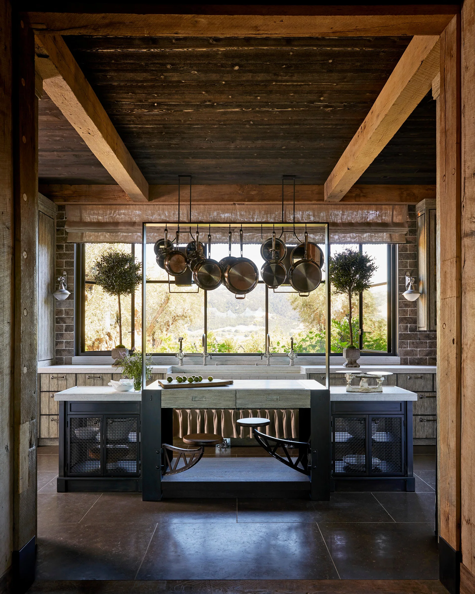 A rustic kitchen features a dark wood ceiling with beams, a central island with a metal pot rack, and a large grid window overlooking a sunny valley.