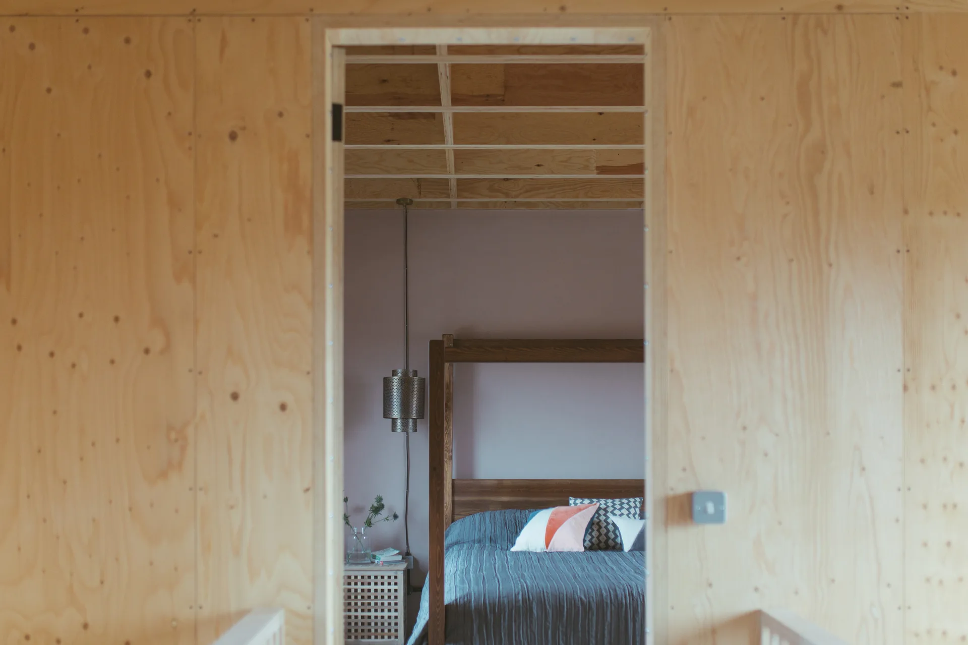 A view through a raw plywood doorway shows a bedroom with a dark wooden bed, a mauve wall, and a wooden grid ceiling.
