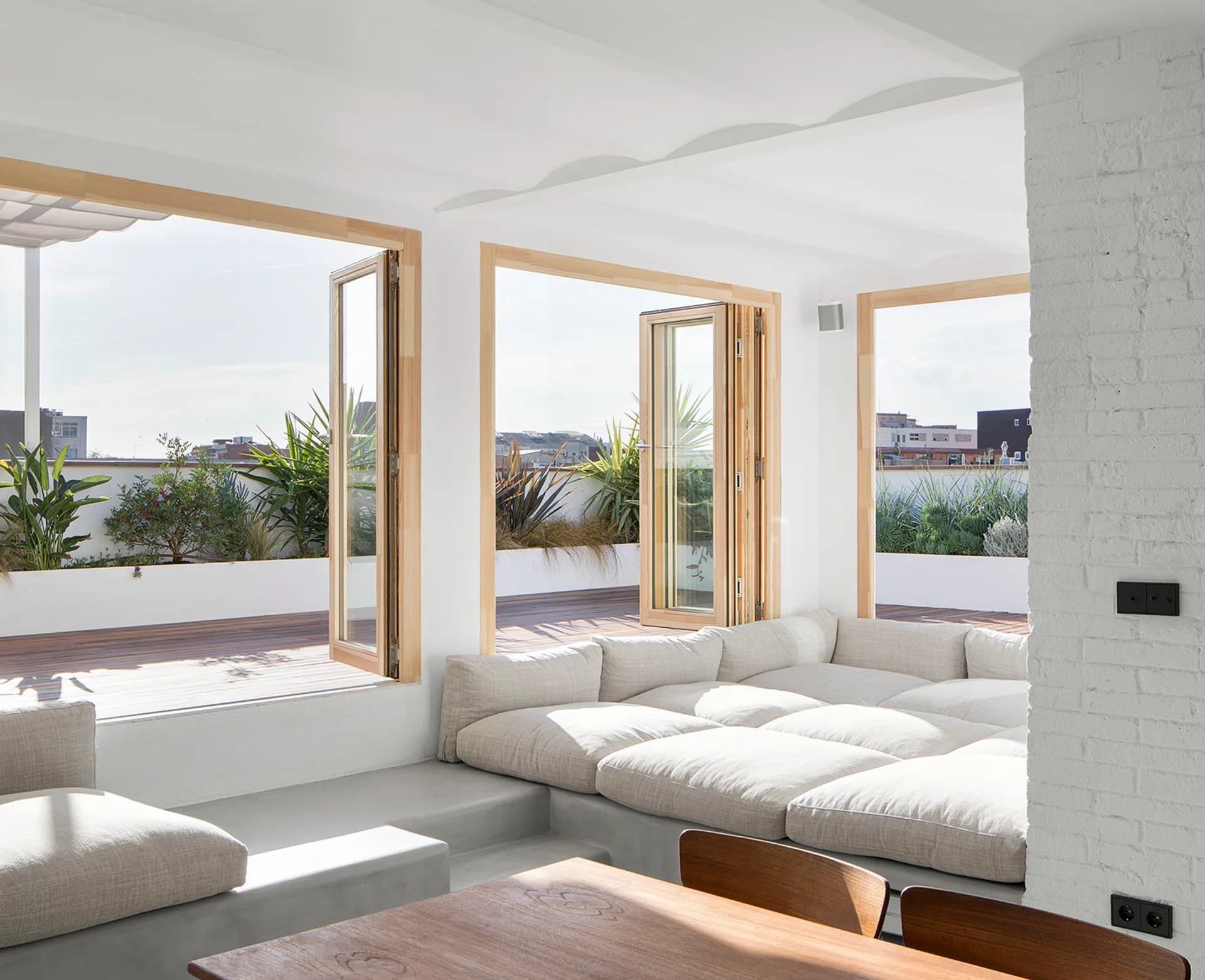 A sunlit living room features a modular beige sofa on a concrete platform next to open wooden bi-fold doors leading to a plant-lined terrace.