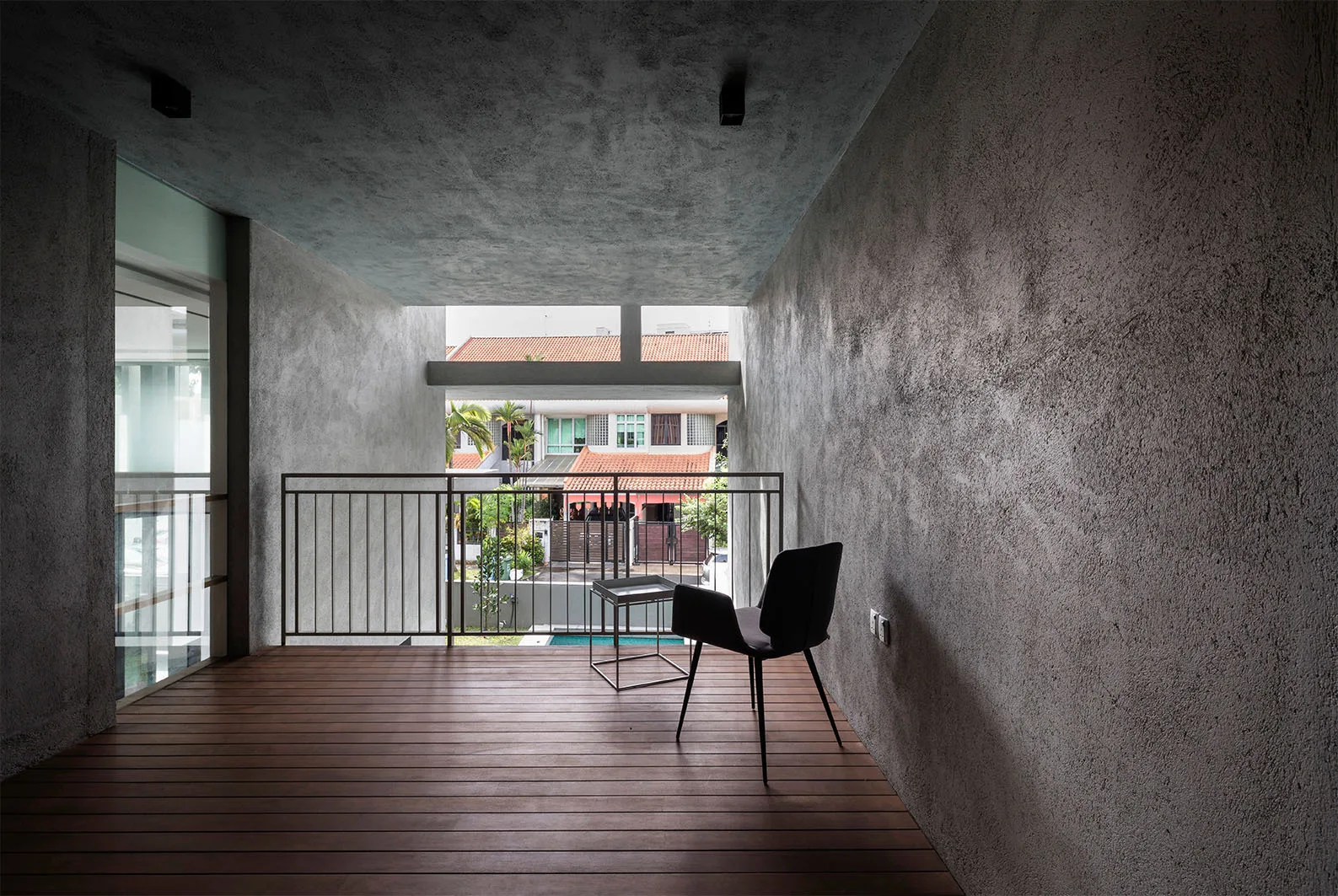 A covered concrete balcony features dark wood plank decking, textured grey walls, a single black chair, and a metal railing looking out over houses.
