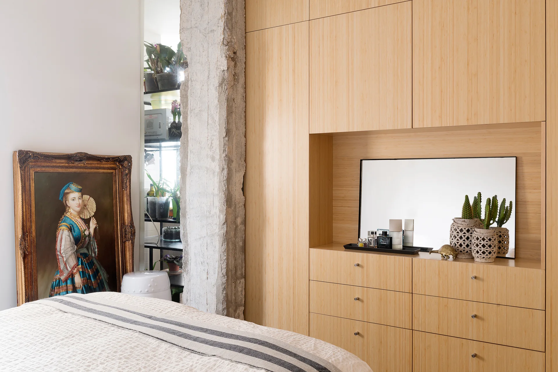 A bedroom features custom light wood built-in cabinetry with a recessed vanity niche, a raw concrete column, and a gold-framed portrait on the floor.