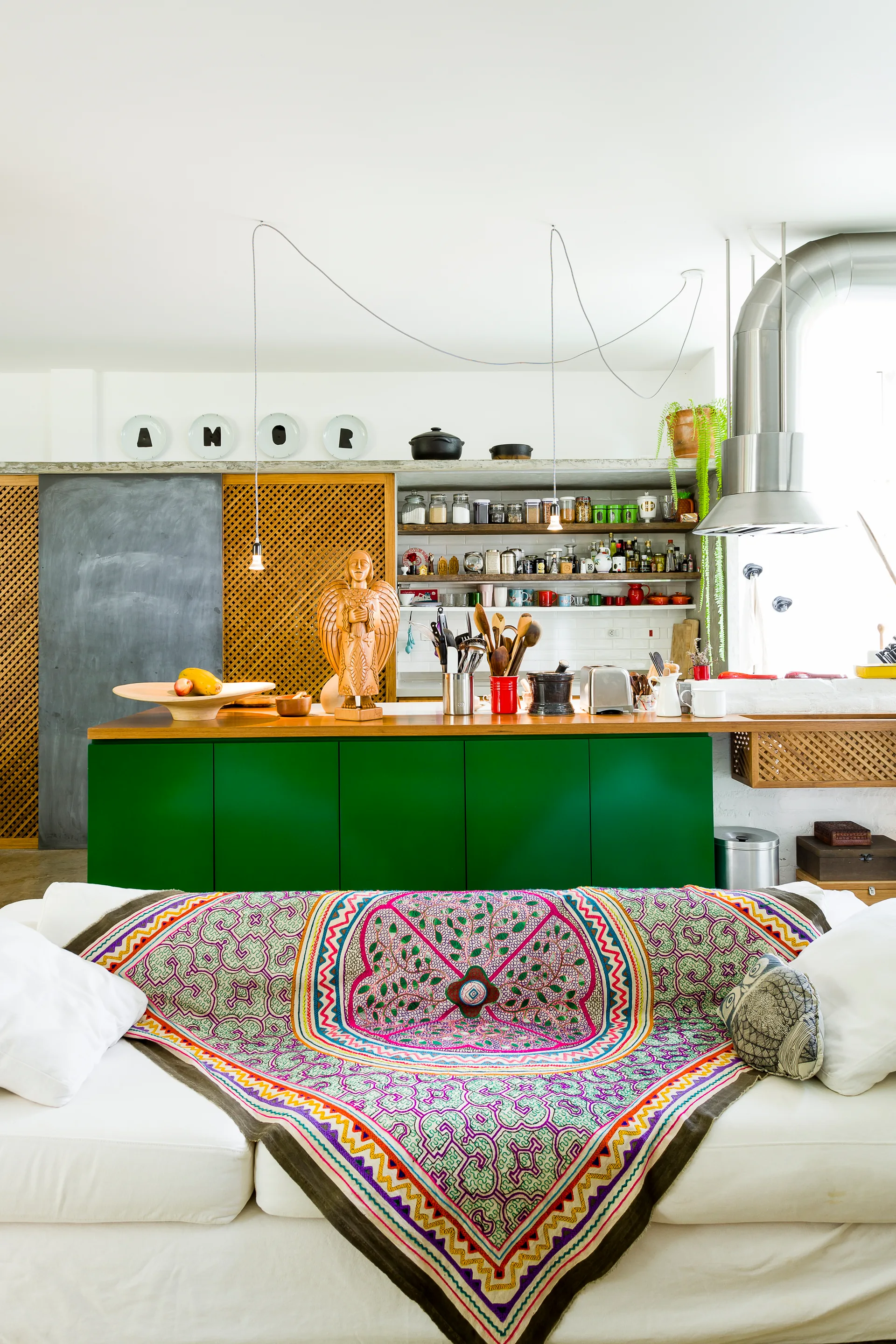 An open-concept kitchen features a bold green island with a wood countertop, open wall shelves, and a white sofa covered in a colorful patterned throw.