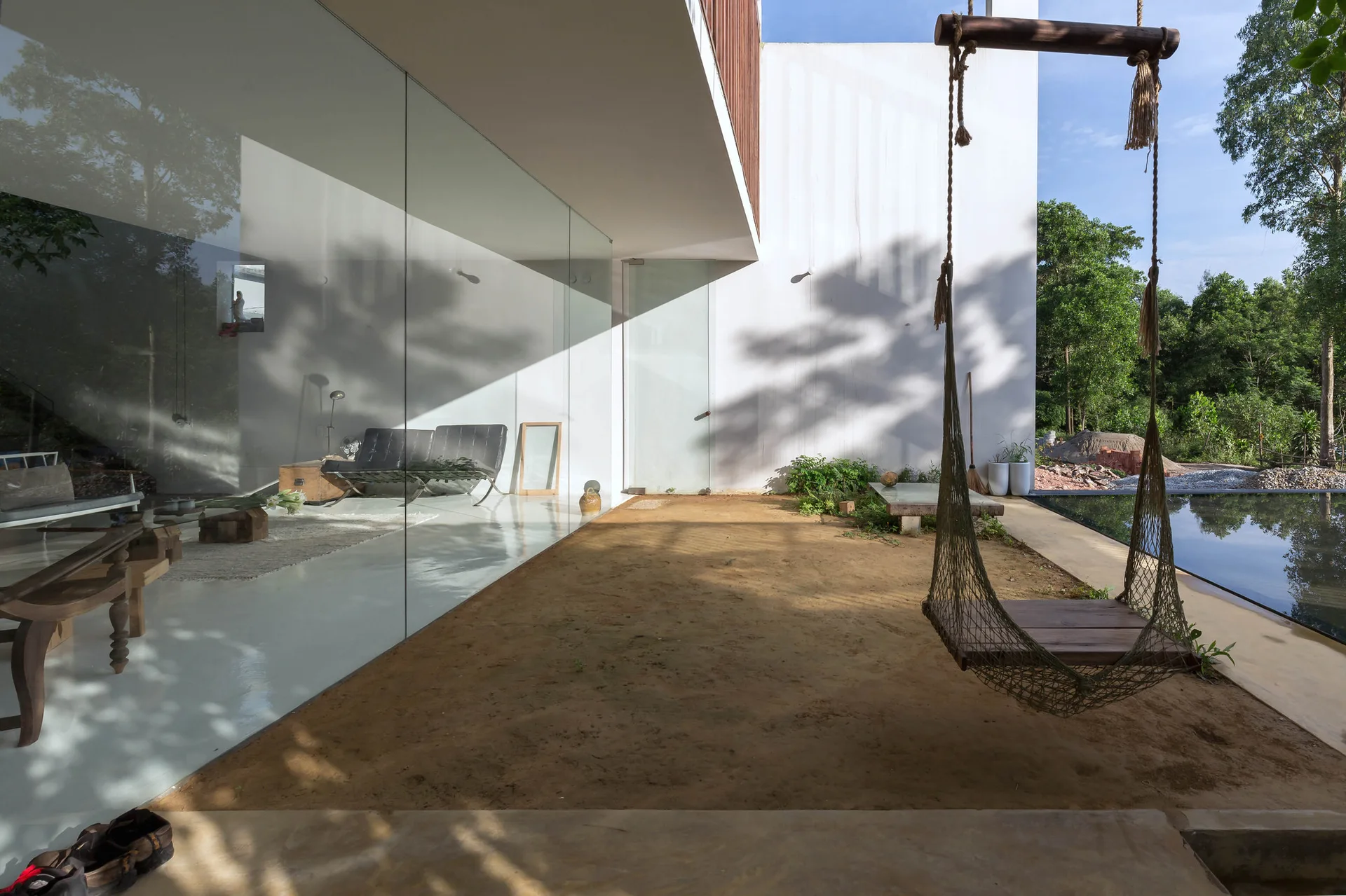 A modern outdoor patio featuring a wooden rope swing, a large glass wall reflecting the interior, and a swimming pool in the background.