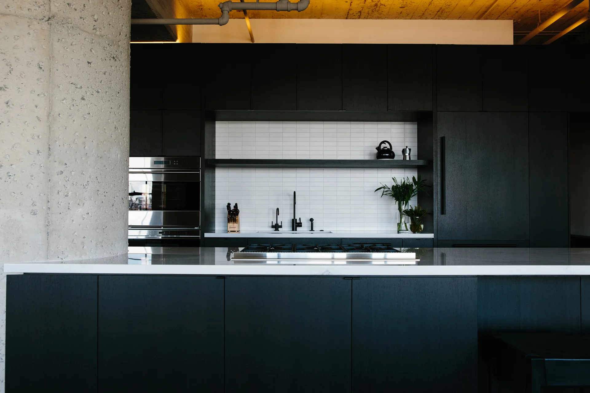 A modern kitchen features matte black cabinets, a white subway tile backsplash, a stone-topped island, and a textured concrete support column.