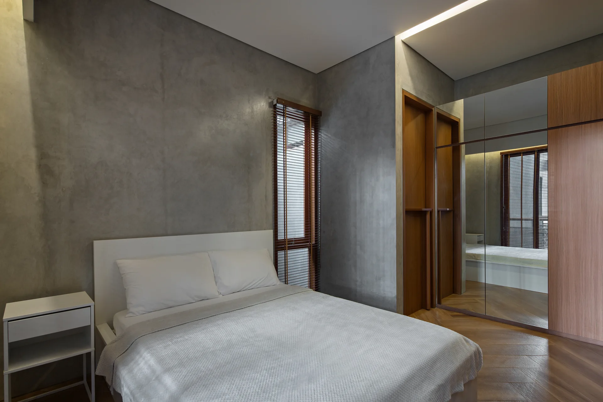 A minimalist bedroom featuring textured gray concrete walls, a white bed, wood flooring, and a mirrored wardrobe.