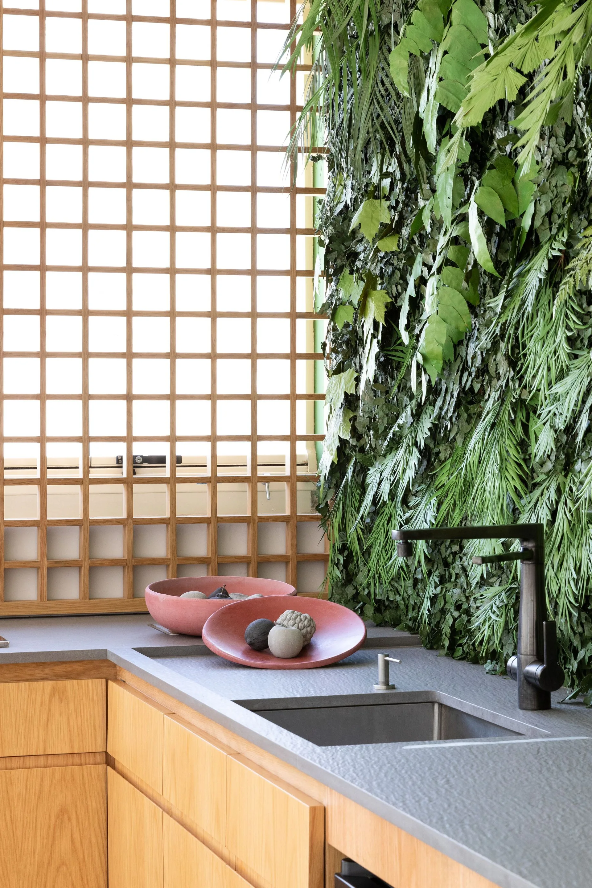 A kitchen sink with a dark faucet, a stone-like countertop, a vertical living wall of green foliage, and a wooden lattice window screen.