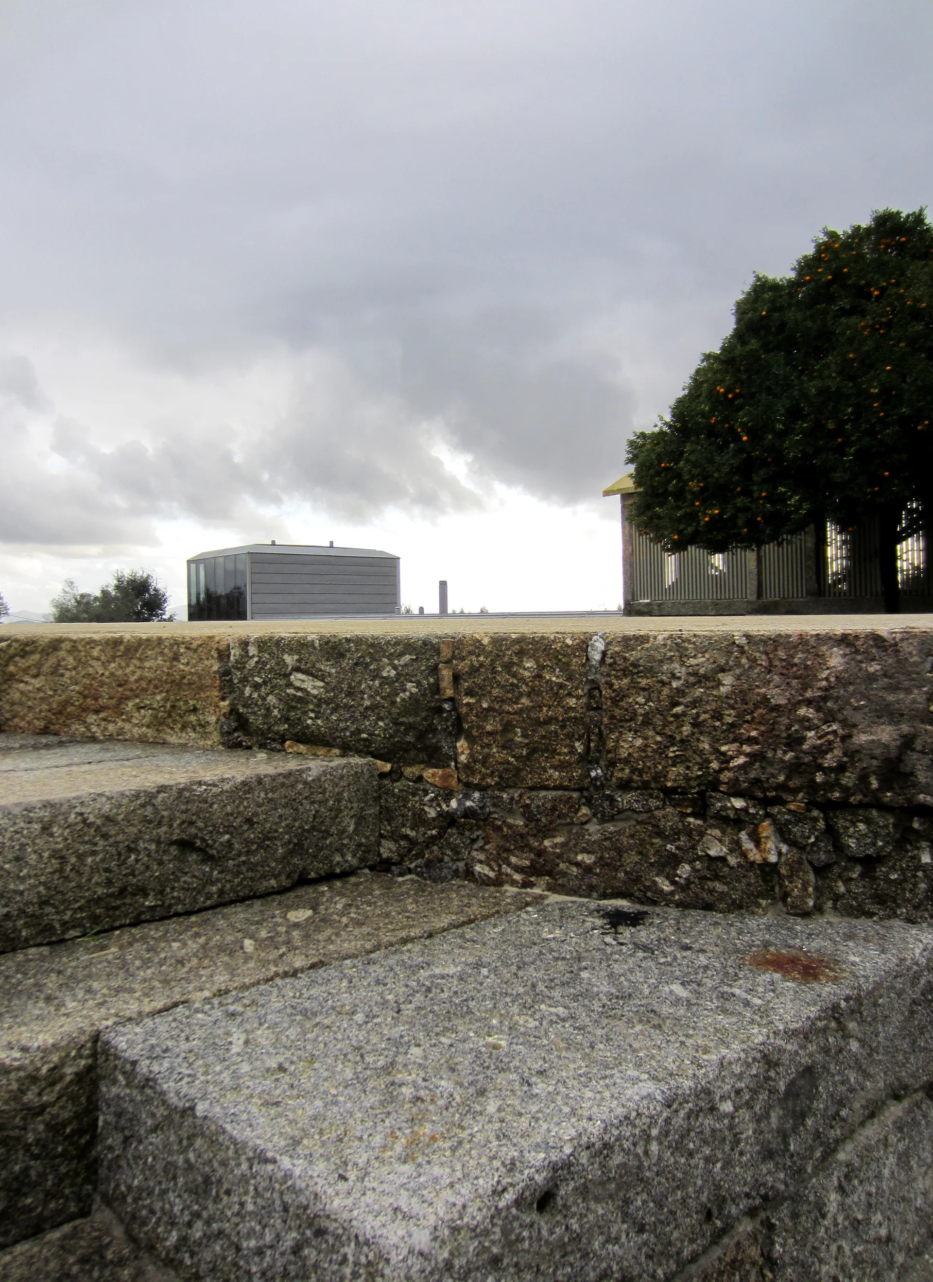 Rough-hewn granite steps lead up next to a stacked stone wall toward an orange tree and a modern gray building under a cloudy sky.