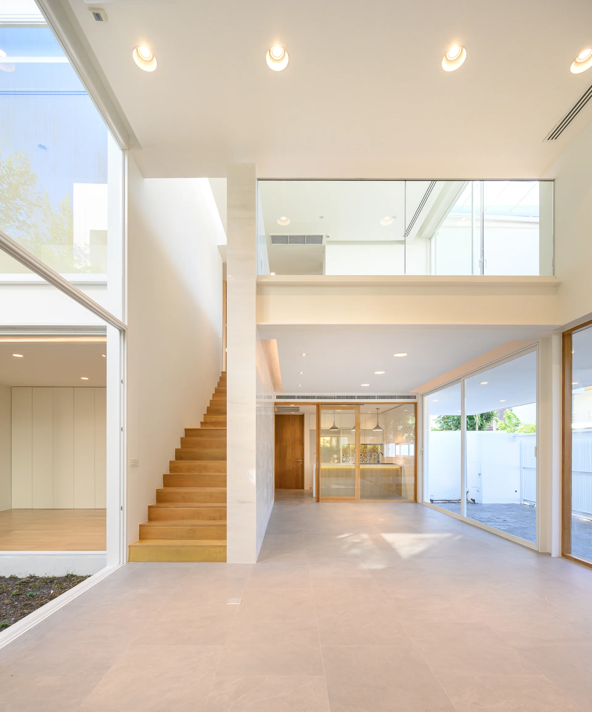 A modern open-plan living space features a light wood staircase, large glass windows, and a glass-railed mezzanine balcony.