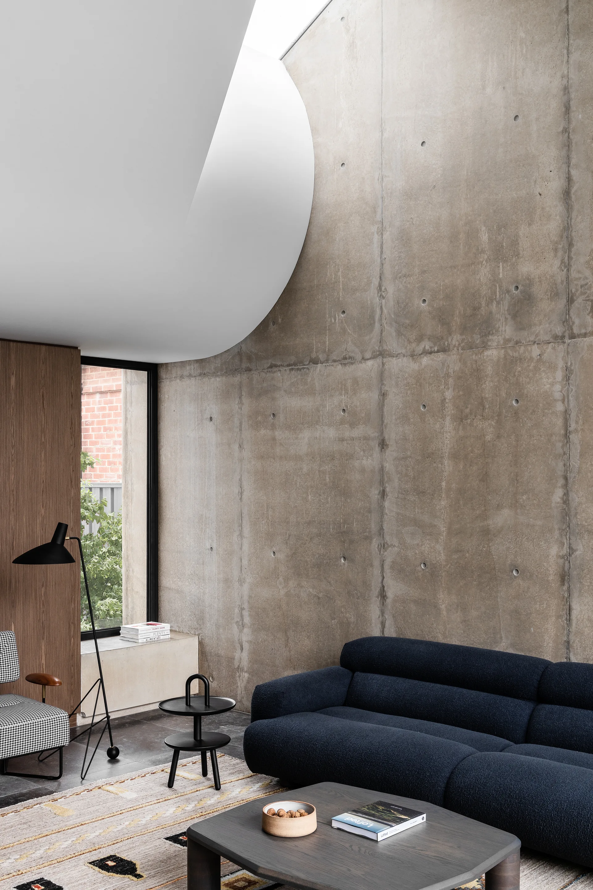 A modern living room features a dark blue textured sofa and a houndstooth armchair against a tall exposed concrete wall with a curved ceiling cutout.