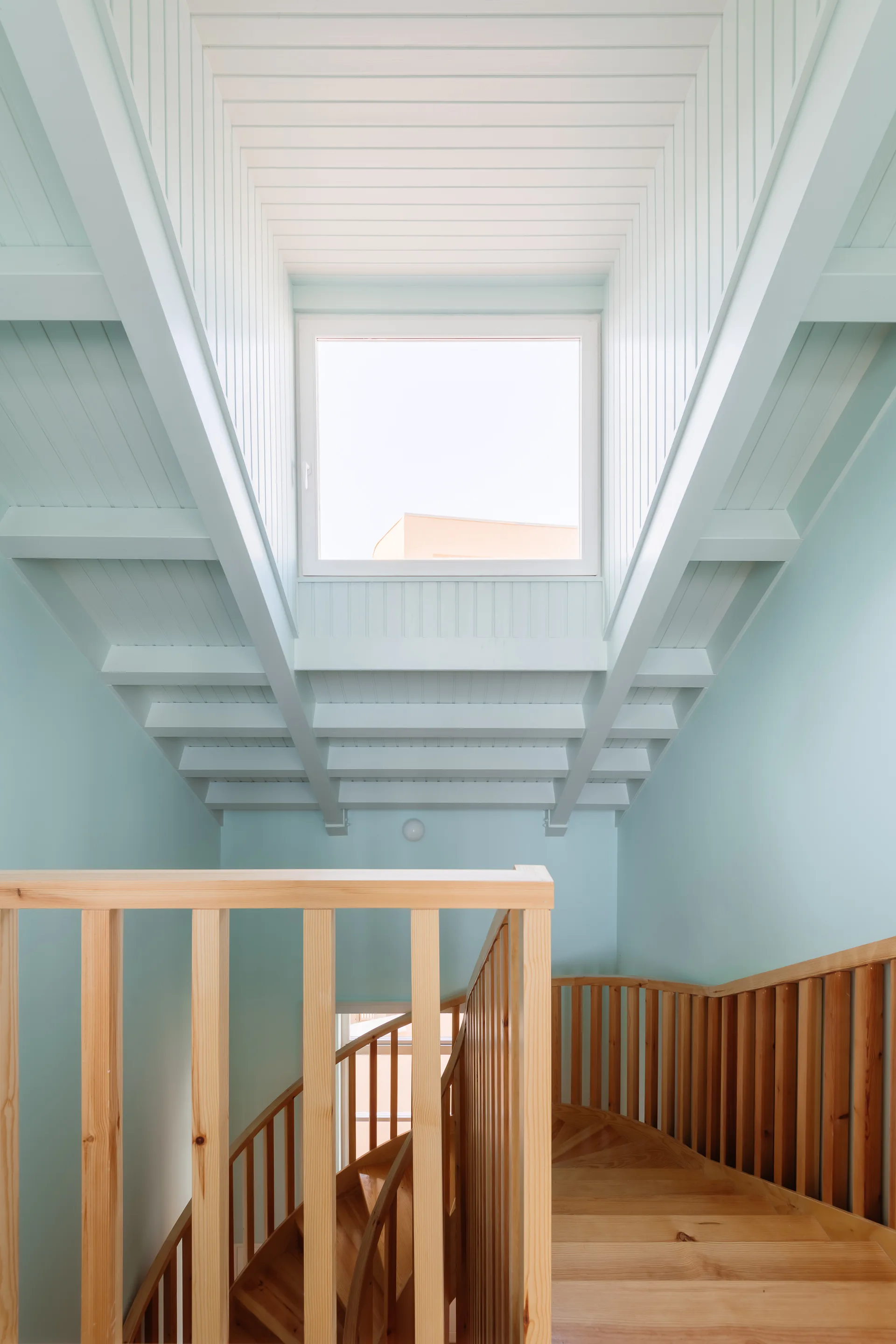 A light wood spiral staircase ascends toward a white paneled ceiling with a square skylight, flanked by pale blue walls.