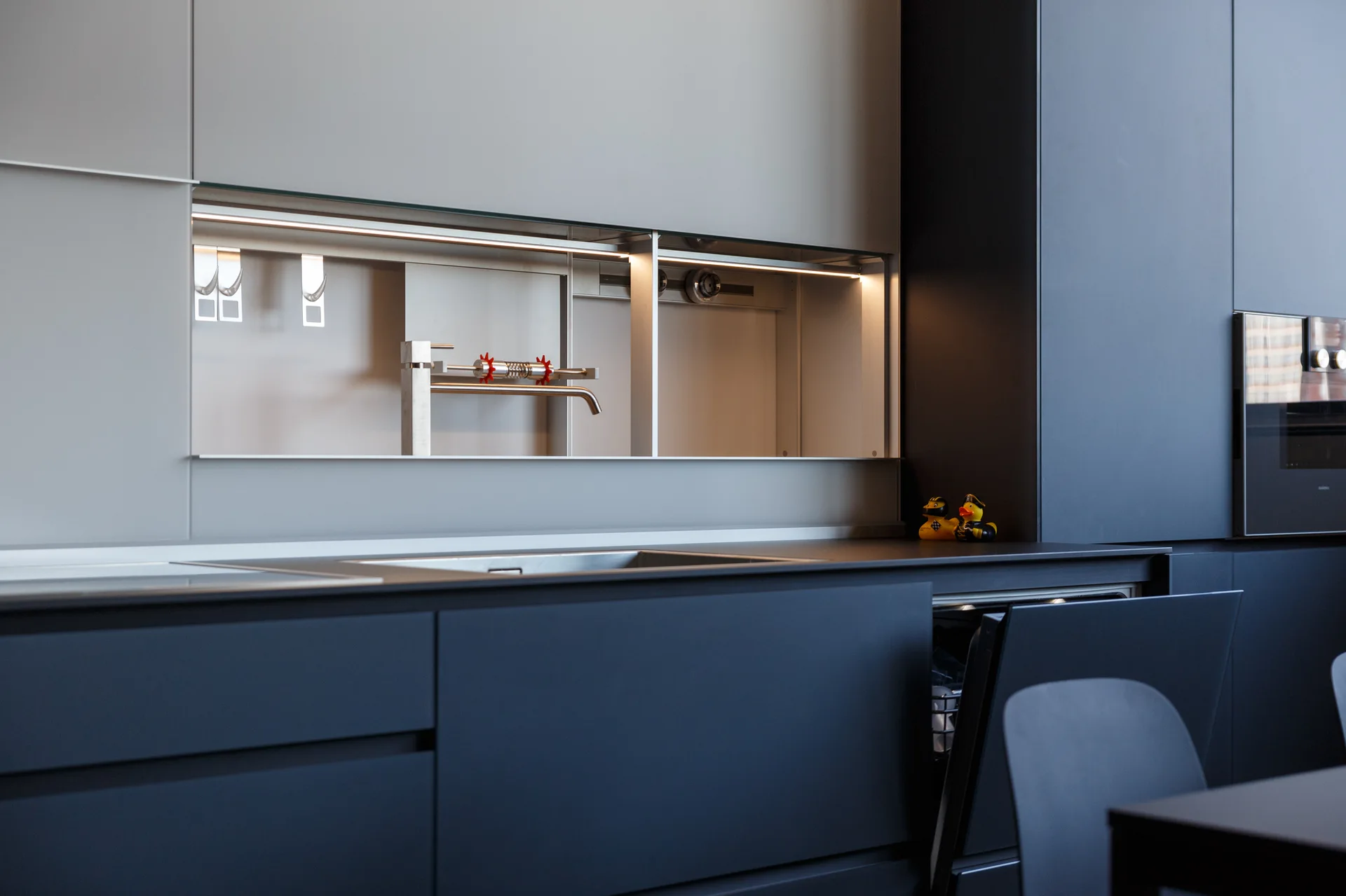 A modern minimalist kitchen with matte black lower cabinets, grey upper cabinets, an integrated sink, and an illuminated backsplash niche.