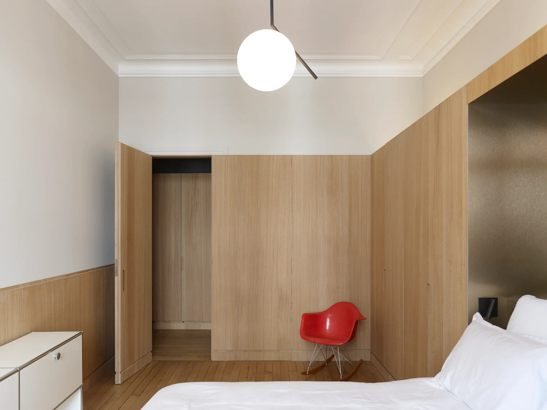 A minimalist bedroom features integrated light wood cabinetry, a white globe pendant light, and a vibrant red molded plastic rocking chair.