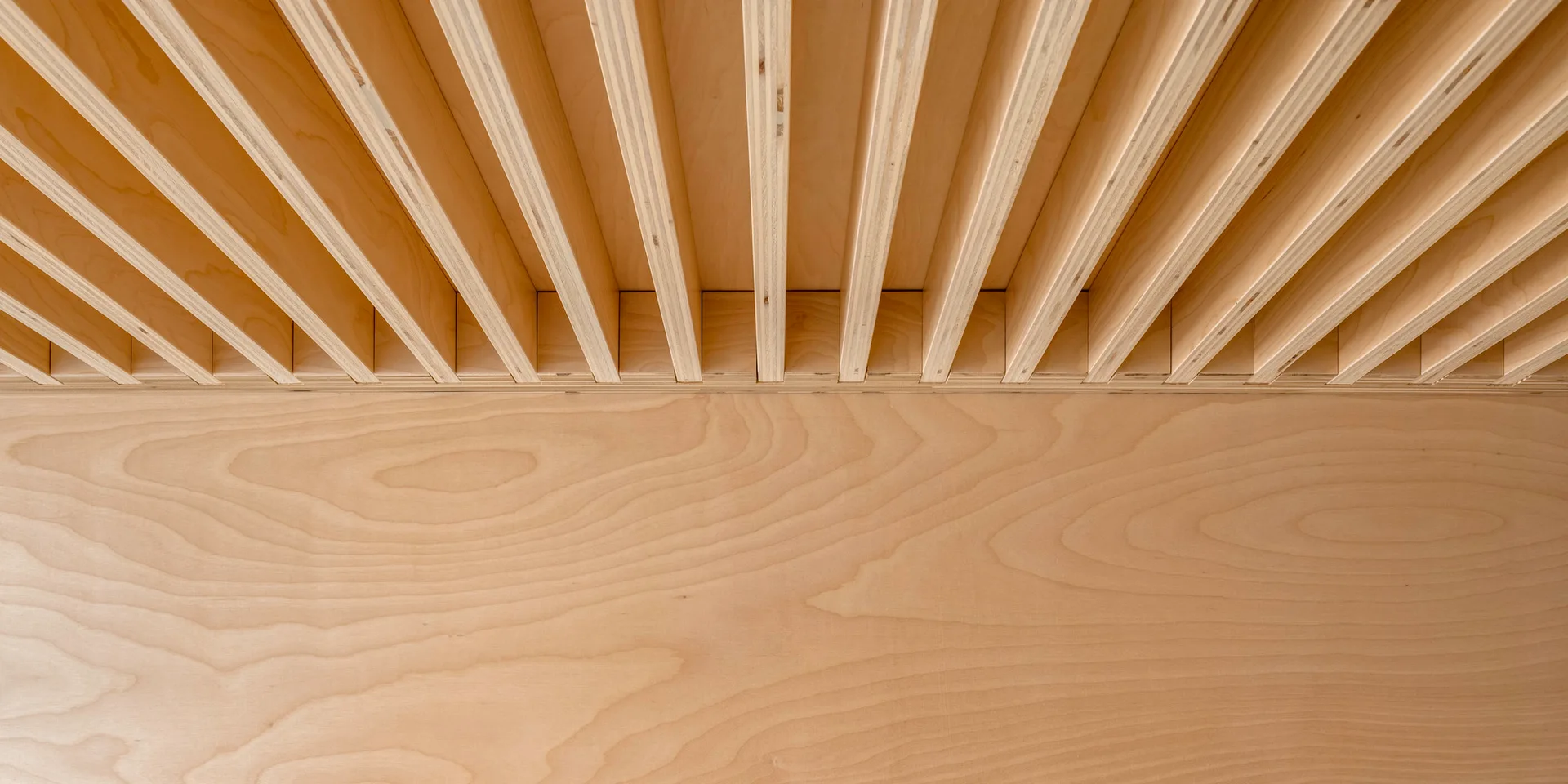 A close-up view of a light-colored wood slatted ceiling meeting a smooth wood veneer wall showing natural grain patterns.