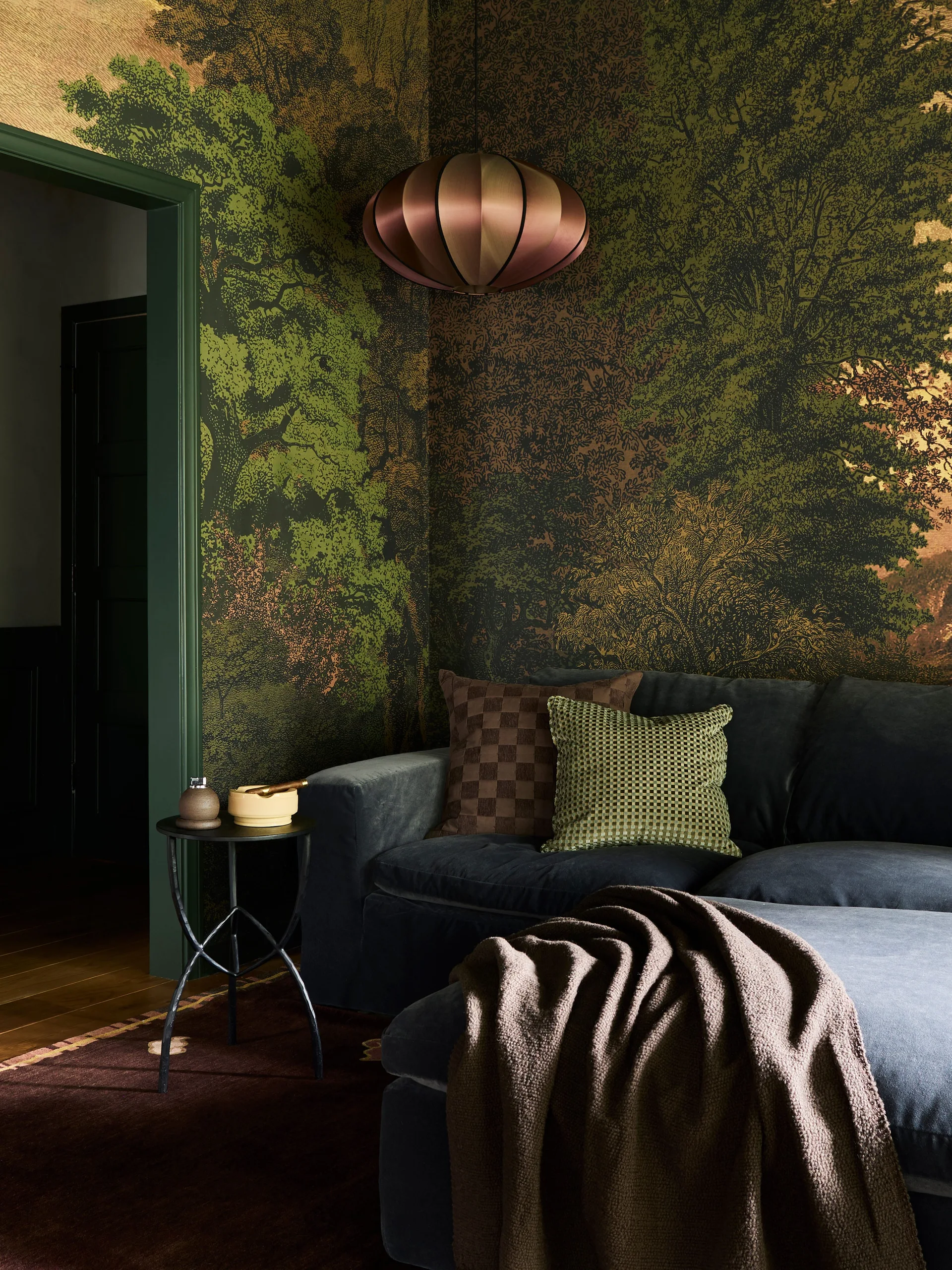 A dark blue velvet sofa sits against a forest landscape mural wallpaper next to a round metal side table.