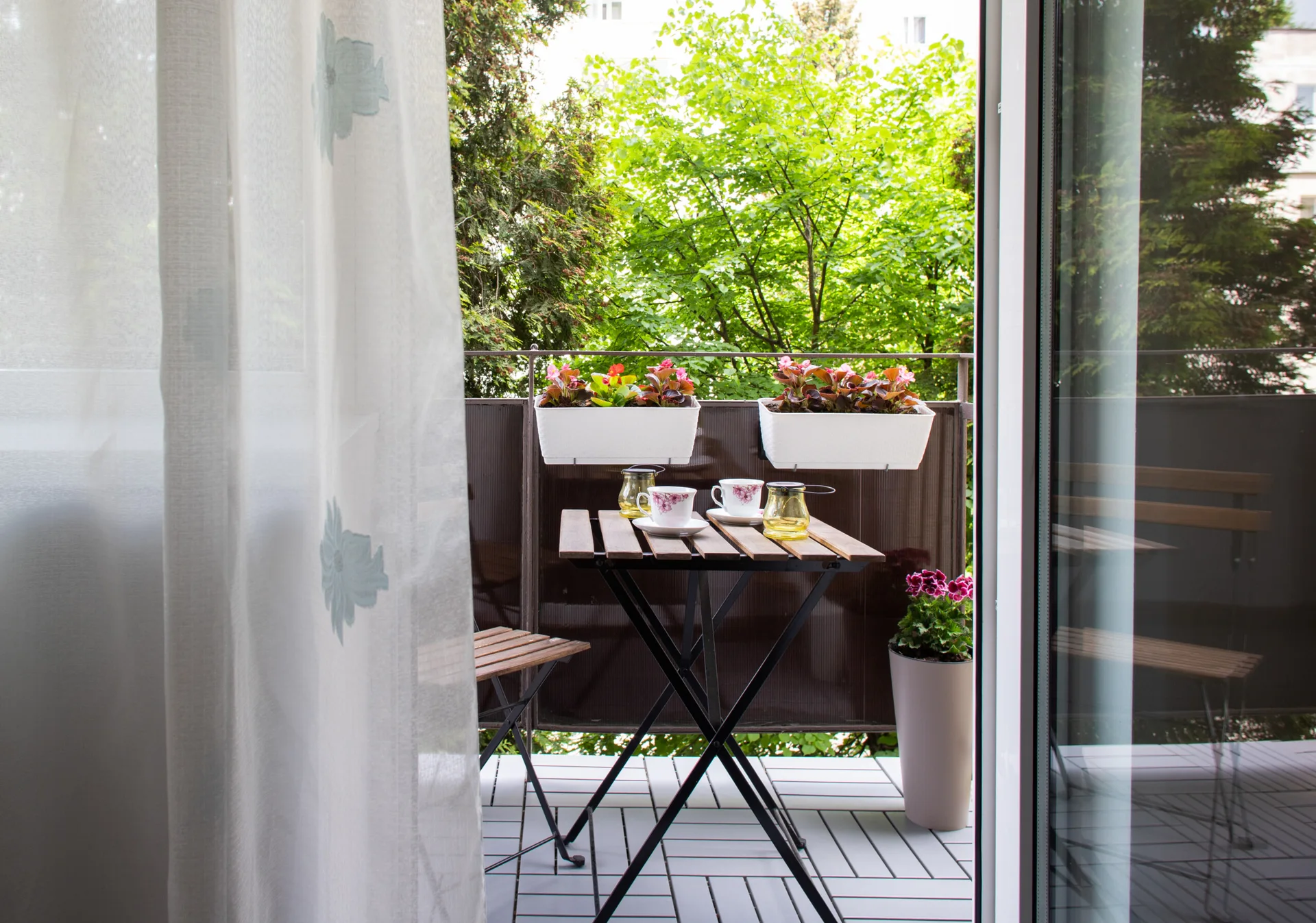 A small apartment balcony features a wooden folding bistro set, grey floor tiles, and white planters on the railing overlooking green trees.