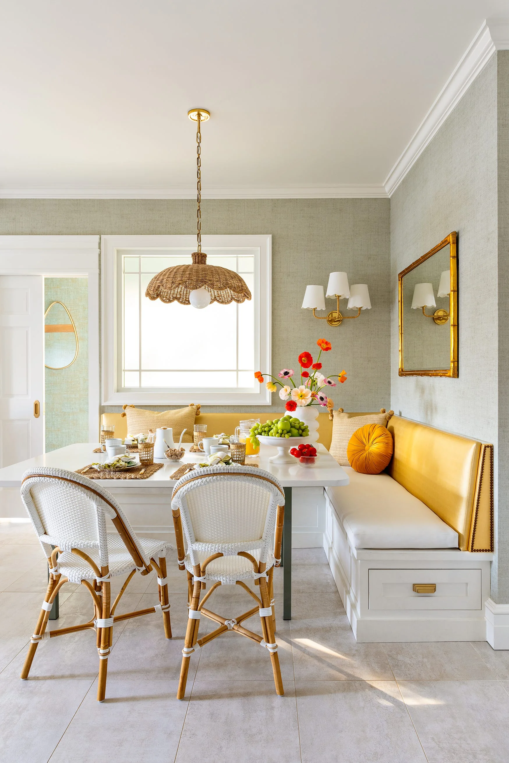 A breakfast nook features a built-in white and yellow banquette, a white dining table, two woven bistro chairs, and a wicker pendant light.