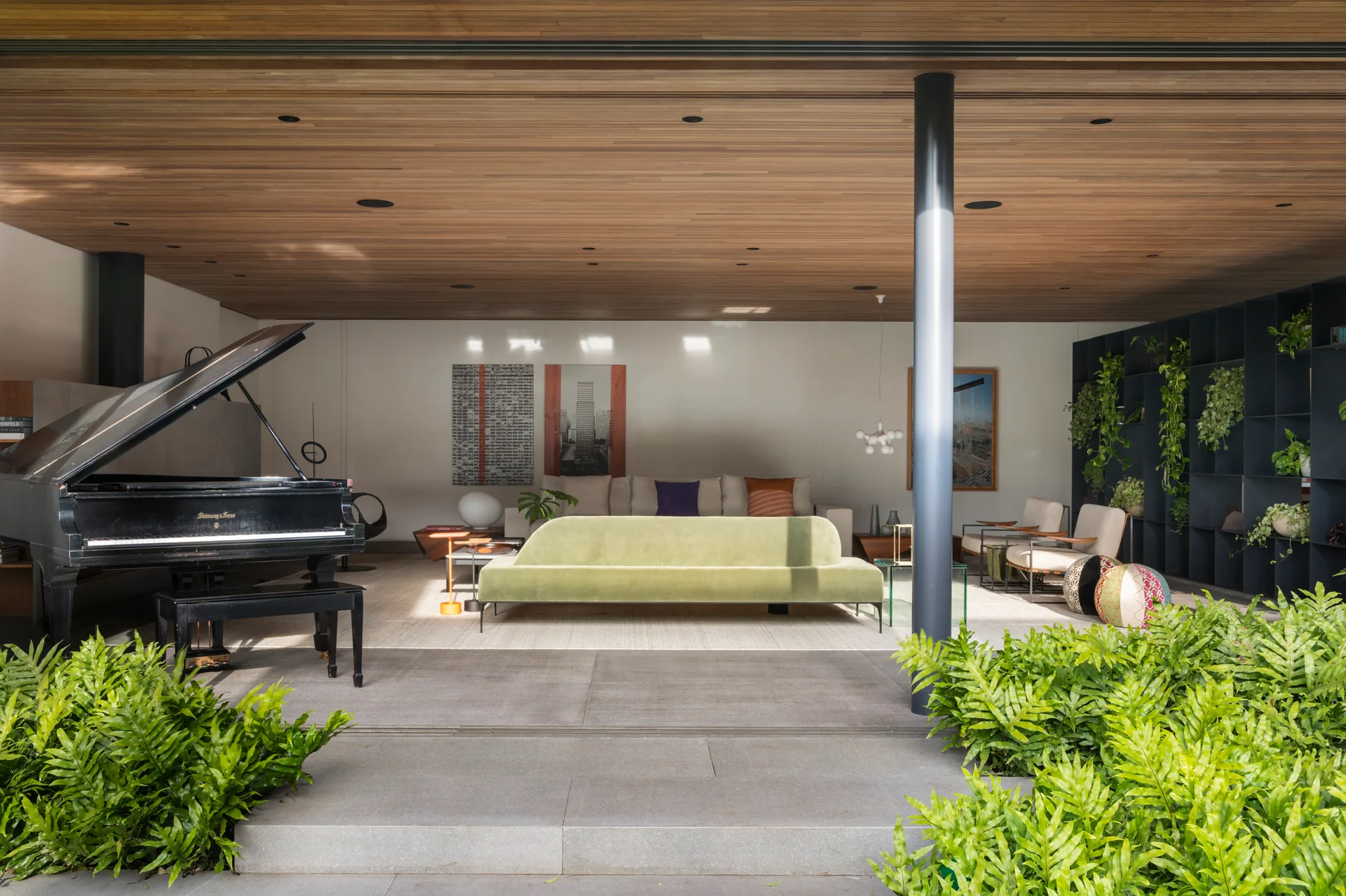 An open living space features a green sofa, black grand piano, and a wood-paneled ceiling that extends over a concrete tile patio.