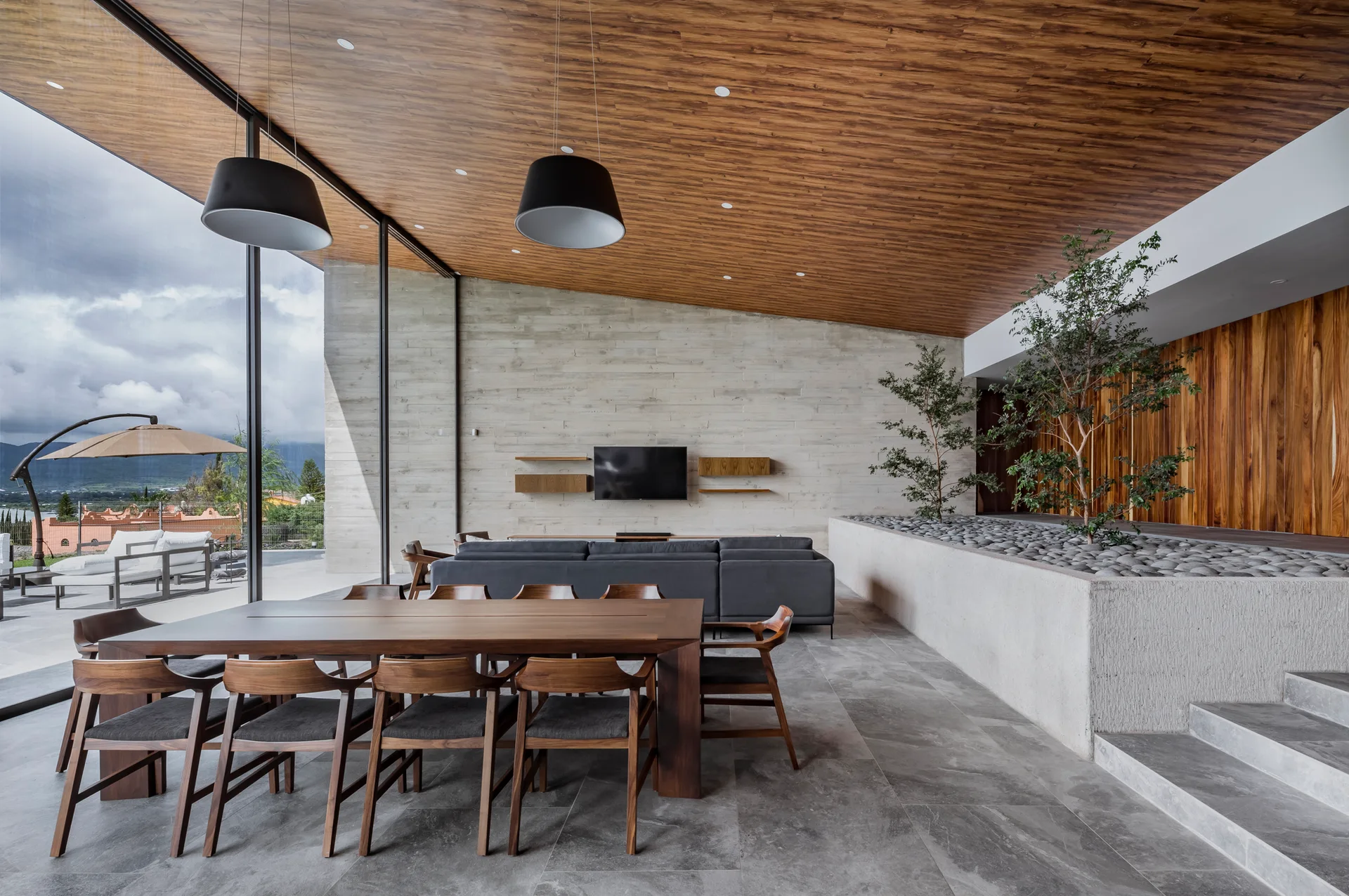 A modern open-plan dining and living room featuring a warm wood plank ceiling, board-formed concrete walls, a large wooden table, and an indoor stone planter.