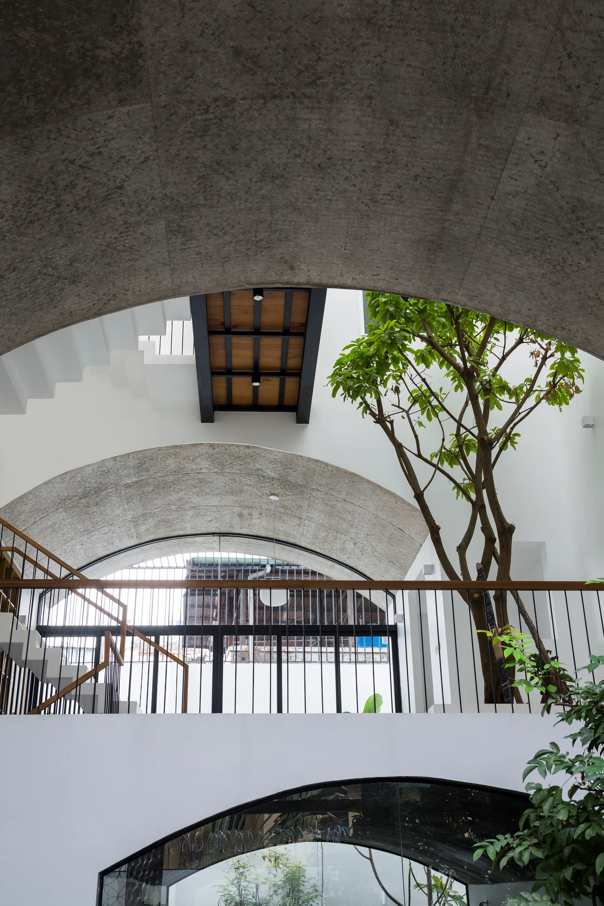 A modern multi-level atrium featuring raw textured concrete arches, white floating stairs, black metal railings, and a tall living tree.