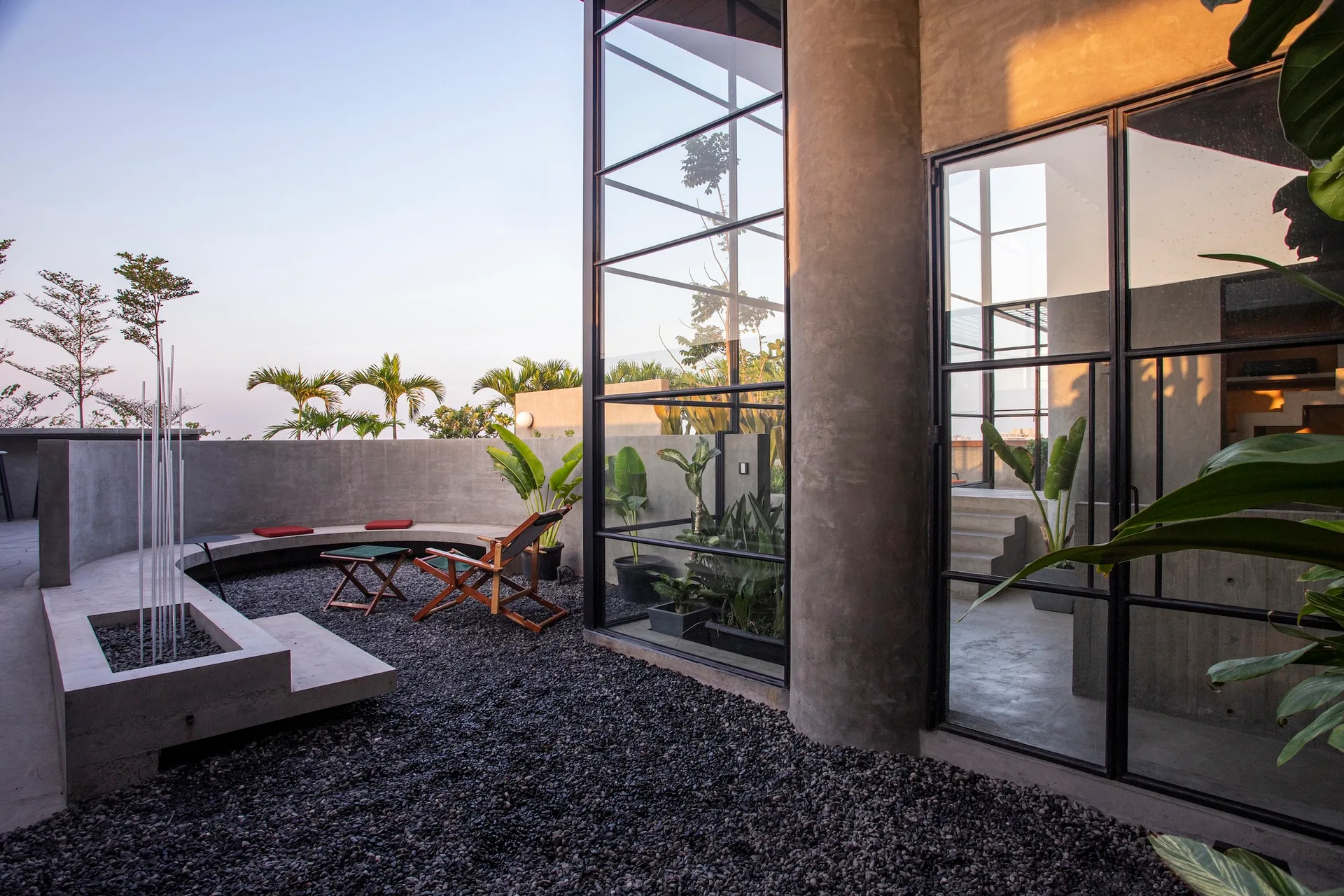 A modern outdoor terrace features a curved concrete wall with integrated seating, dark gravel underfoot, and a large black-framed glass facade.