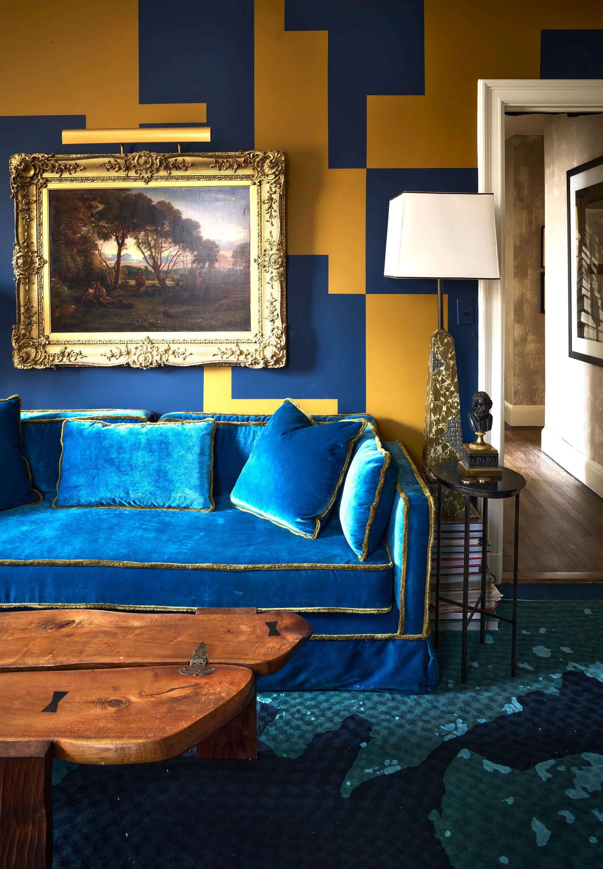 A bright blue velvet sofa sits in front of blue and gold geometric wallpaper beneath a gilded landscape painting.