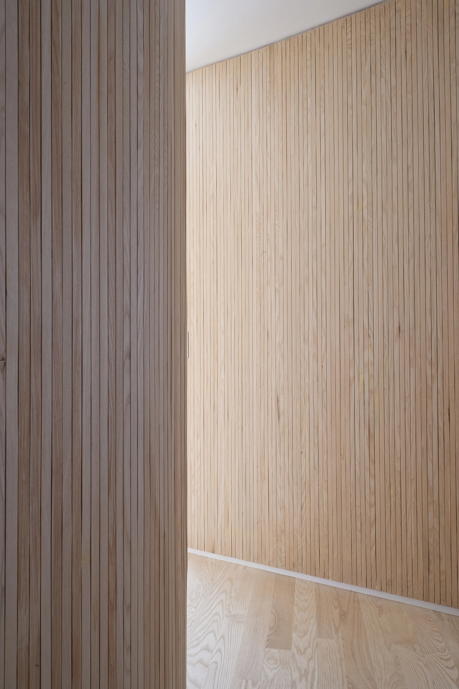 A minimalist hallway features light wood vertical slat wall cladding, matching wood flooring, and an integrated hidden door.