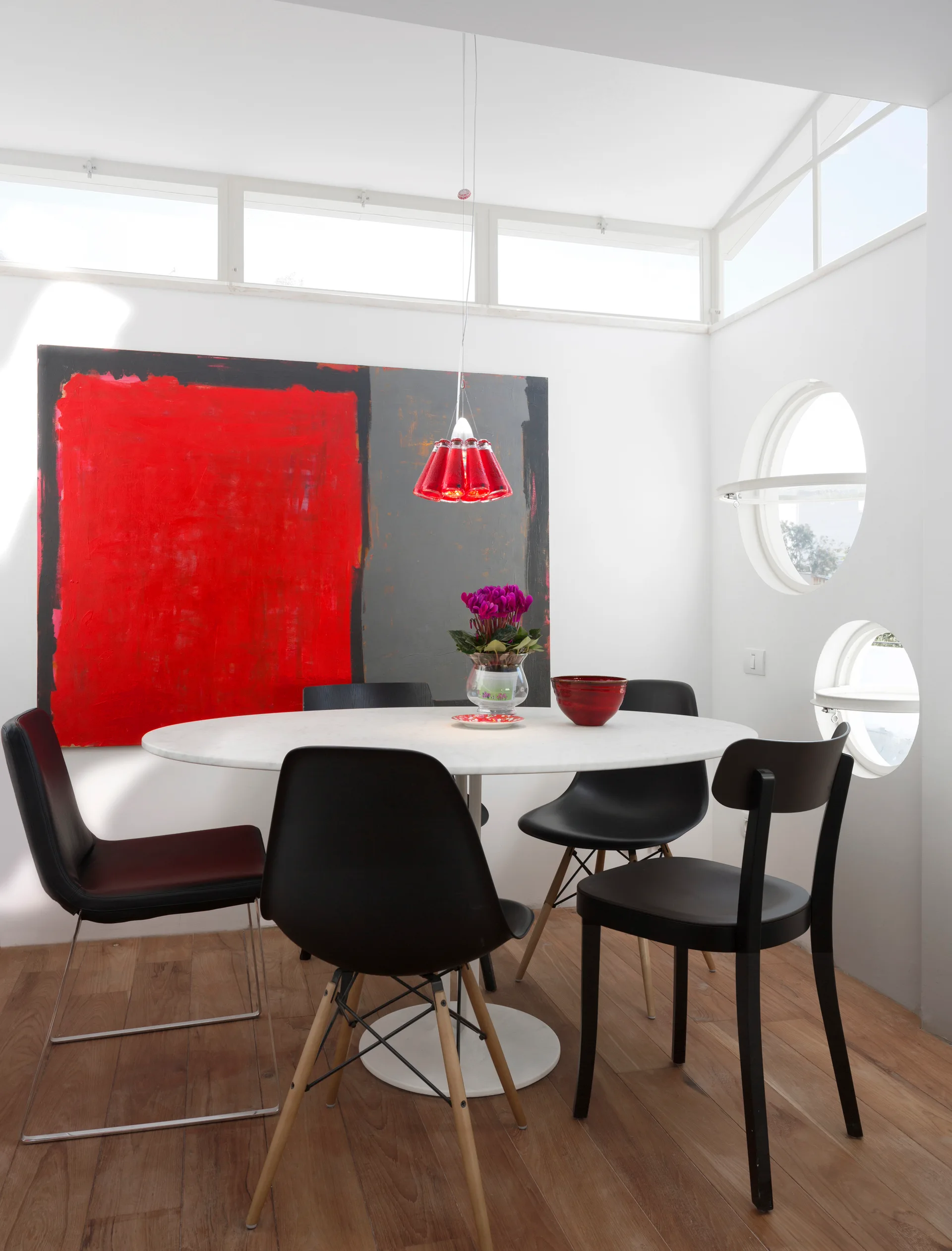 A modern dining room features a round marble table surrounded by black chairs beneath a red pendant light and large red abstract painting.