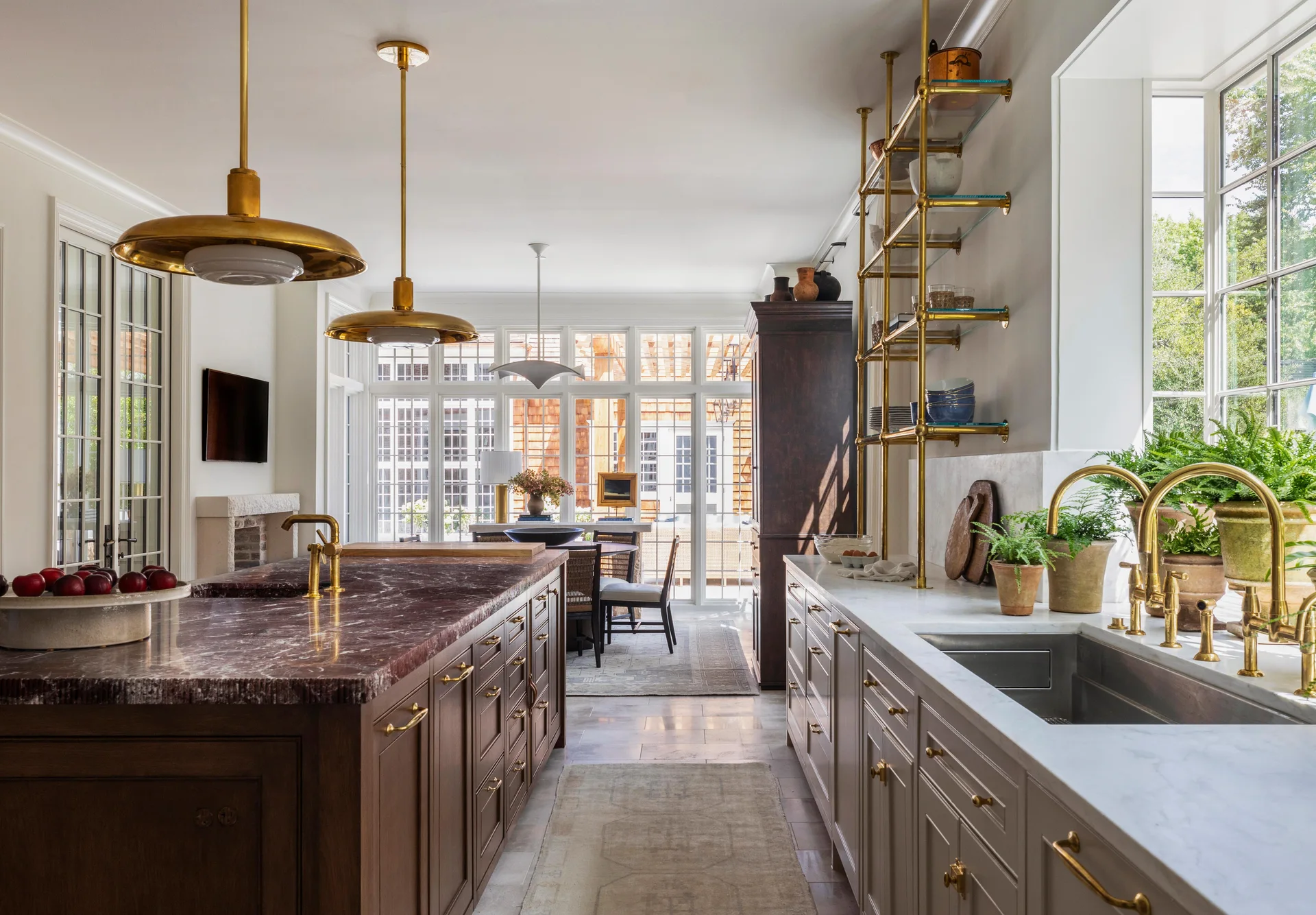 A spacious kitchen features a dark wood island with a veined purple marble top, brass pendant lights, and light gray counter cabinets with brass faucets.