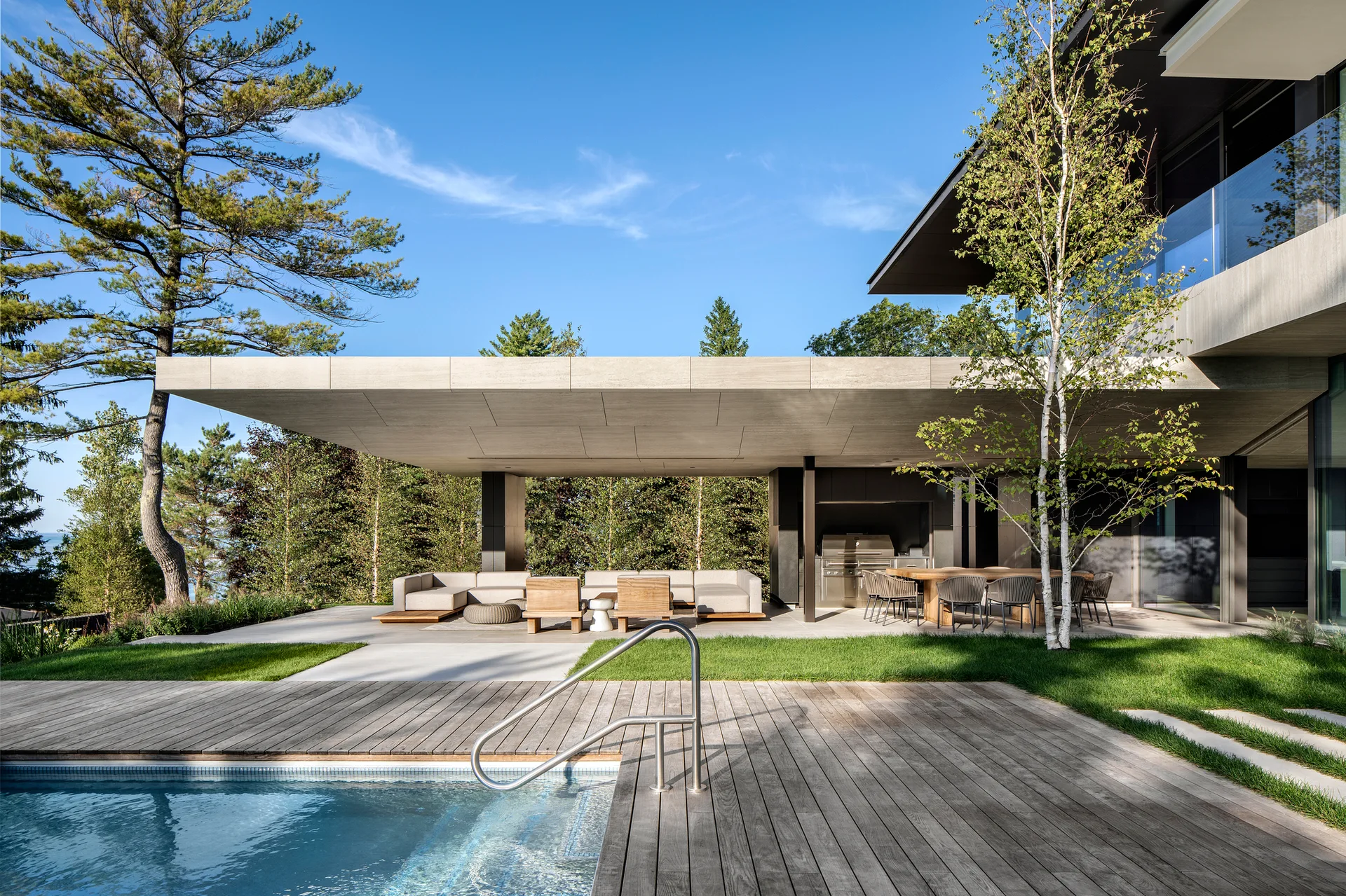 A modern poolside patio features a large cantilevered concrete overhang shading a lounge sectional, outdoor dining table, and built-in kitchen.