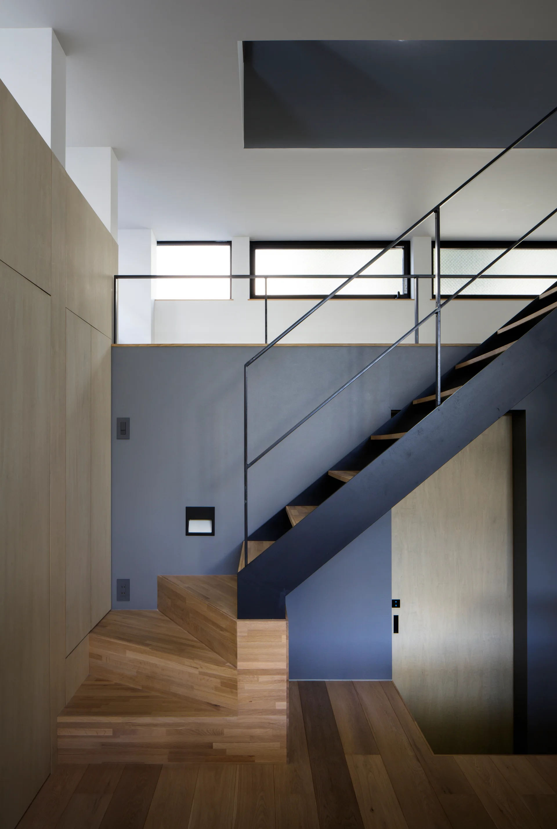A modern interior staircase with light wood treads, a black steel frame and railing, set against a matte gray wall and natural wood paneling.