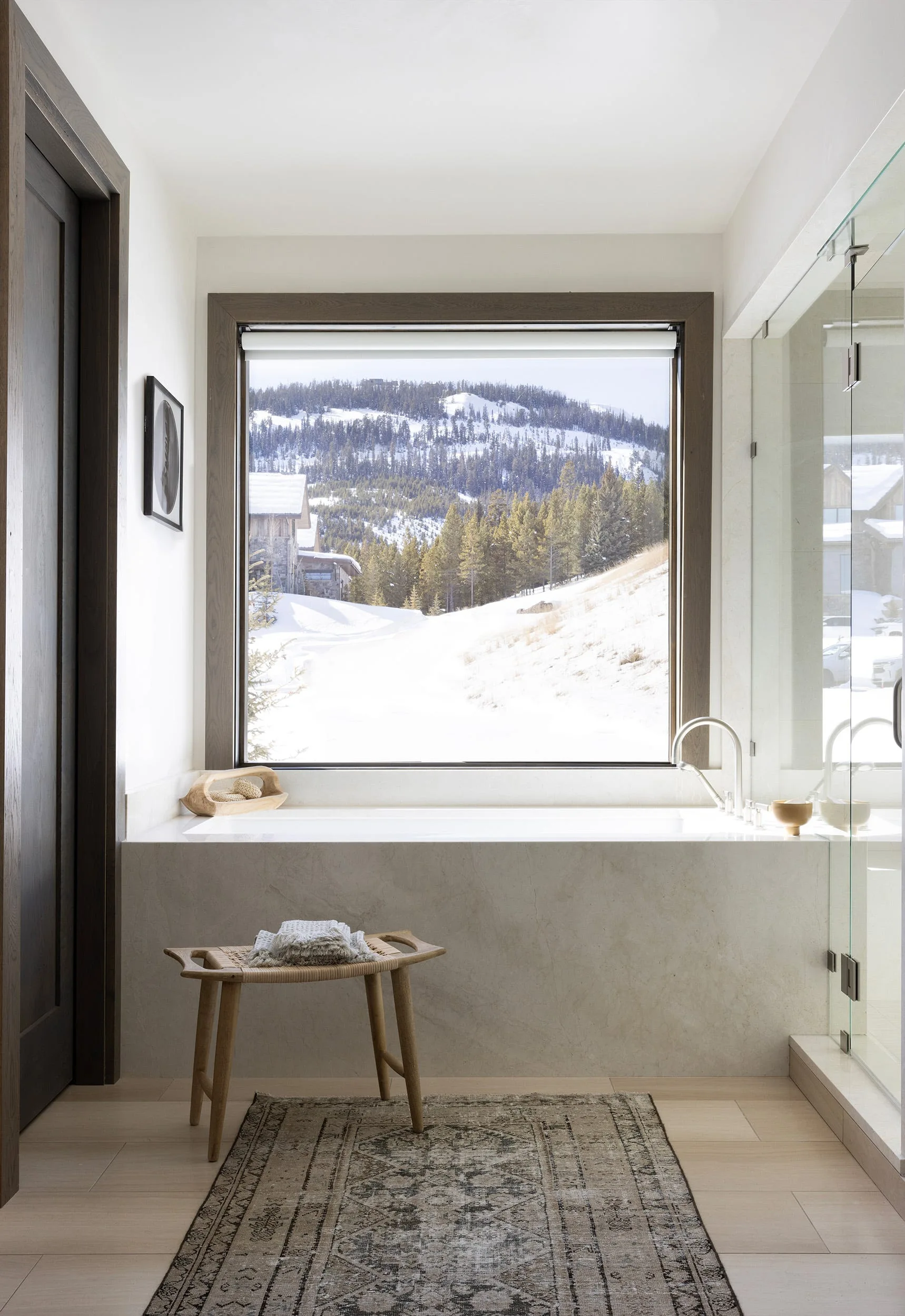A modern bathroom features a stone soaking tub positioned below a large picture window overlooking a snowy mountain pine forest.