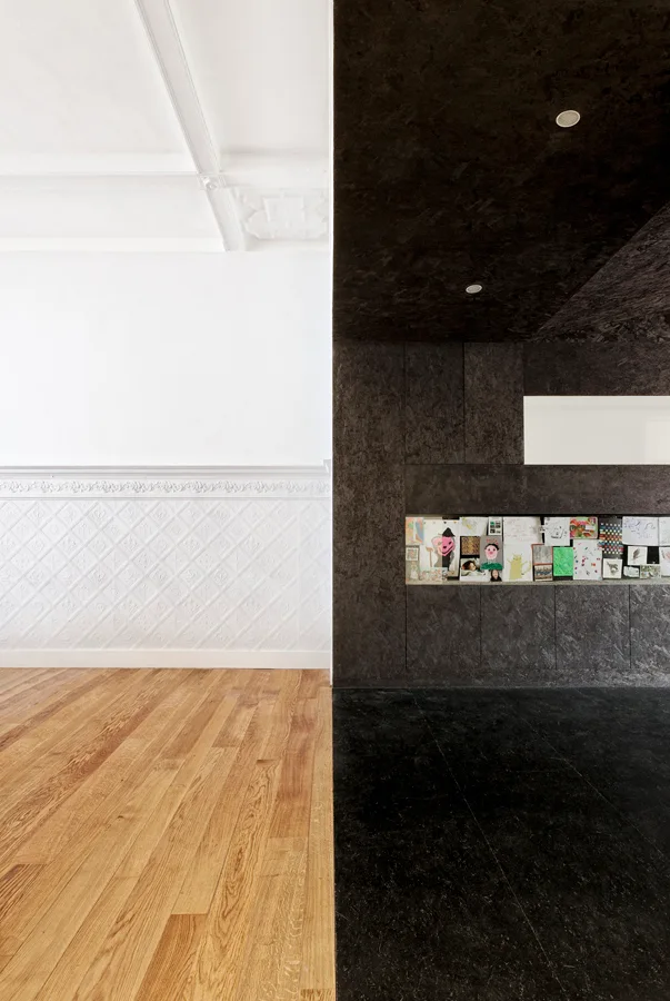An interior wall split vertically between white ornate plaster wainscoting with wood floors and a dark textured OSB paneled wall with black floors.