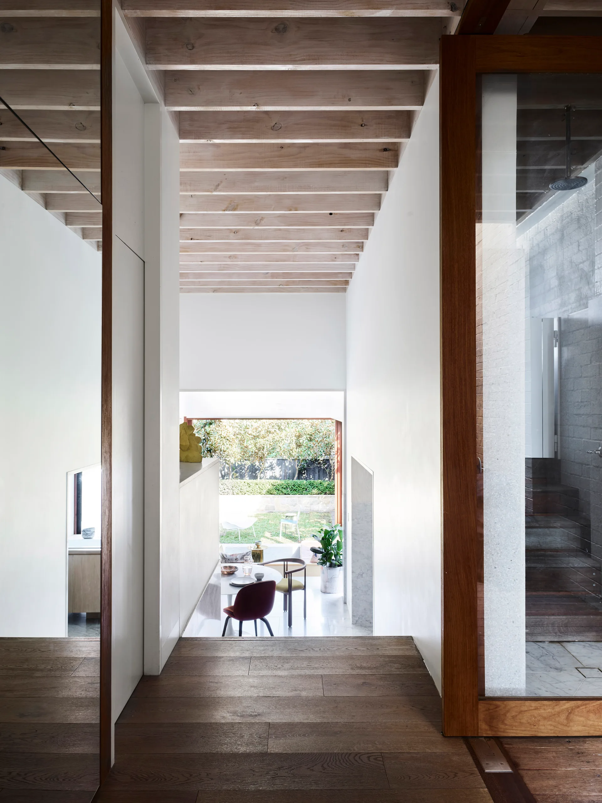 A hallway with dark wood floors and white walls features exposed timber ceiling joists and a view looking down toward a dining area and garden.