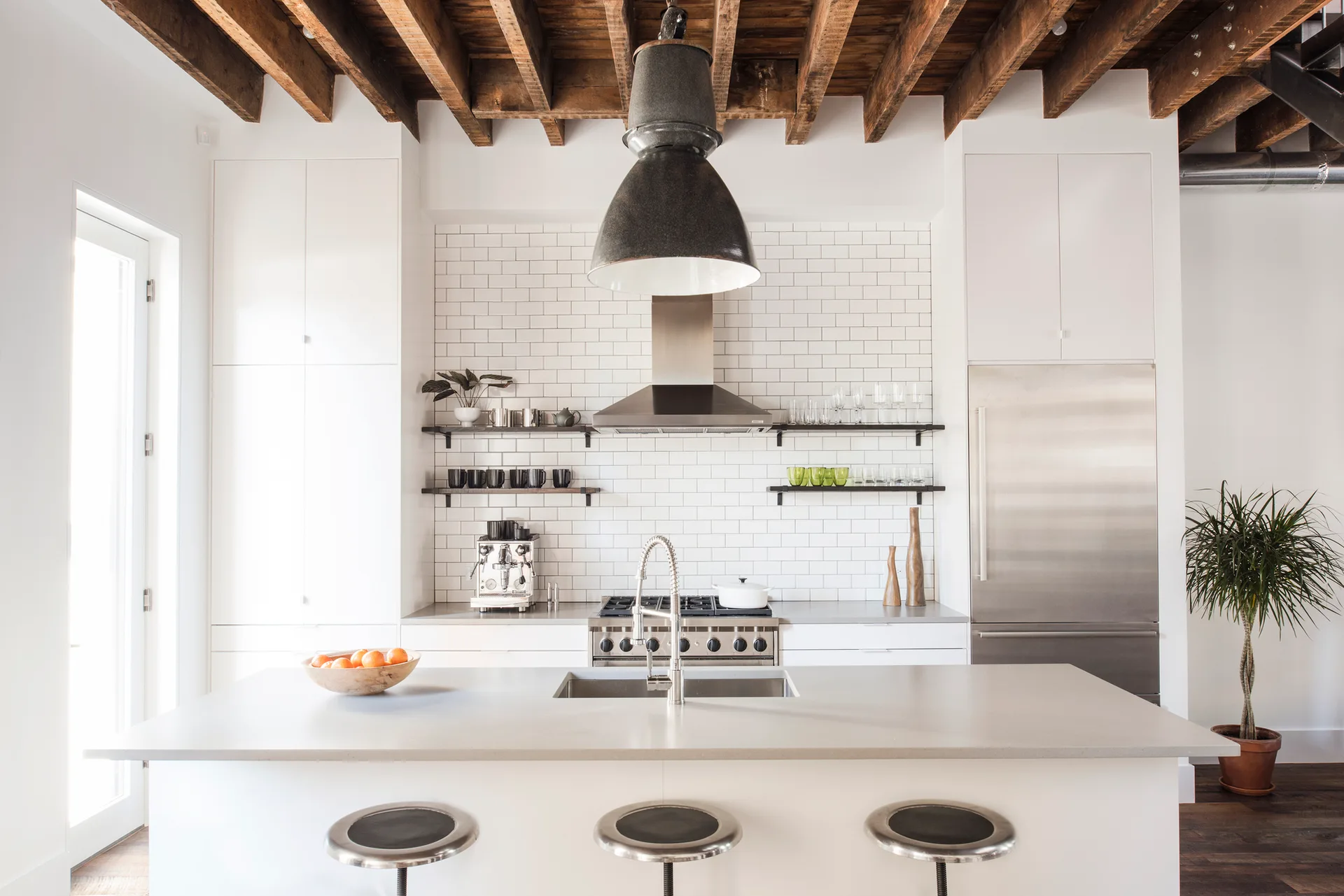 A modern kitchen featuring a central island with three metal stools, white subway tile backsplash, and exposed dark wood ceiling beams.