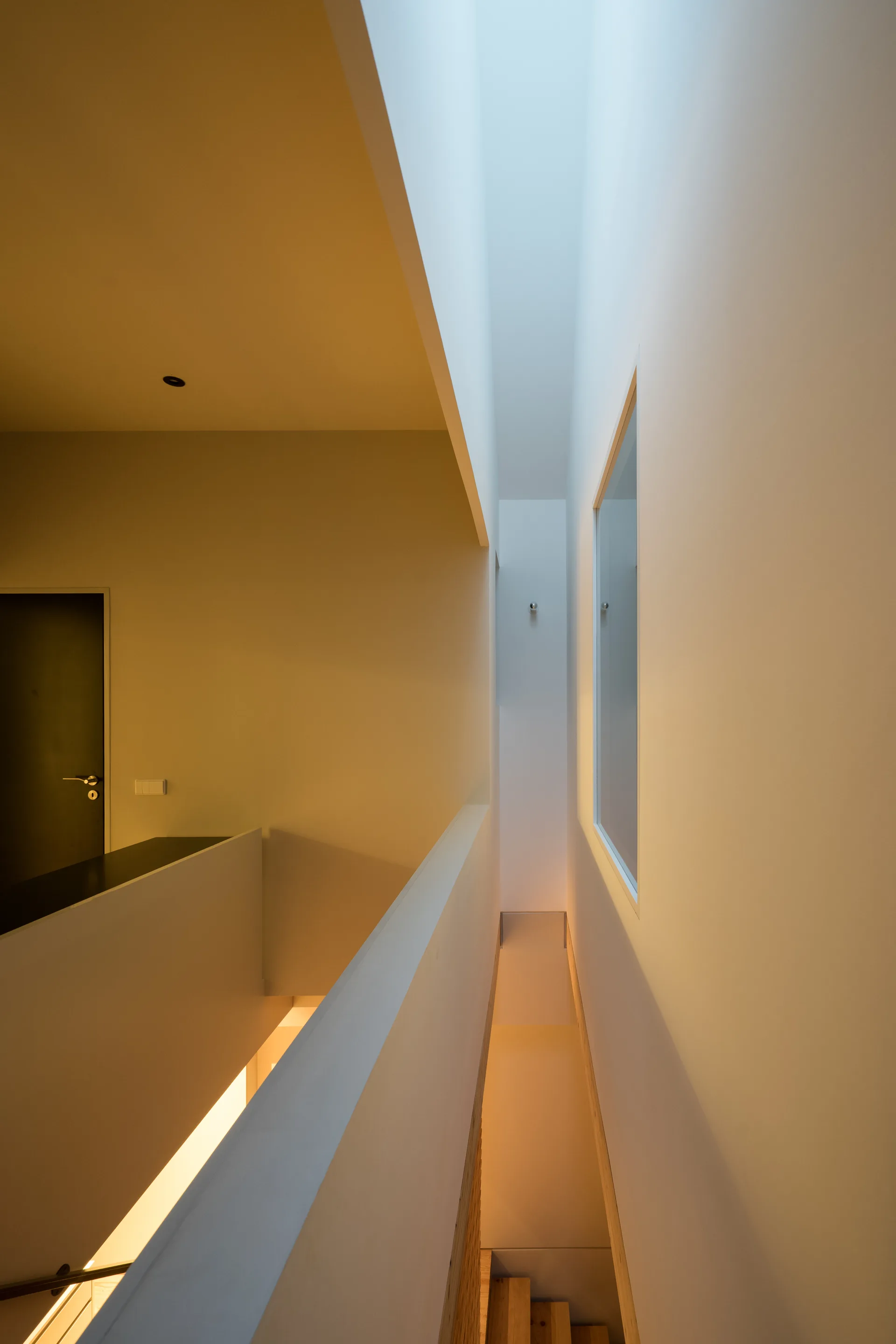 A minimalist white hallway and staircase landing features a long overhead skylight, clean geometric walls, and glimpses of wooden steps below.